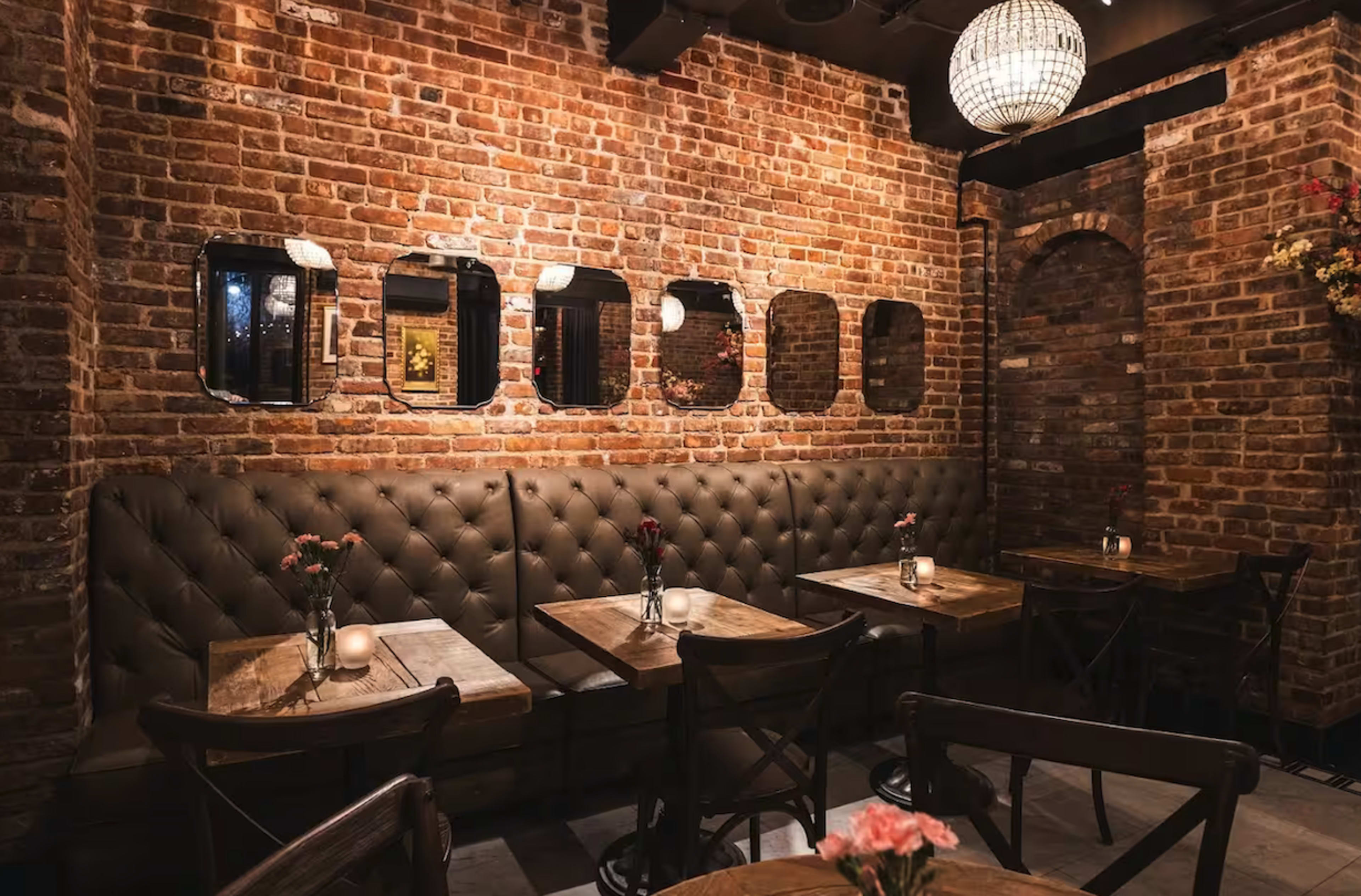 The interior of a restaurant features exposed brick walls, tufted leather seating, and wooden tables with small vases of flowers.