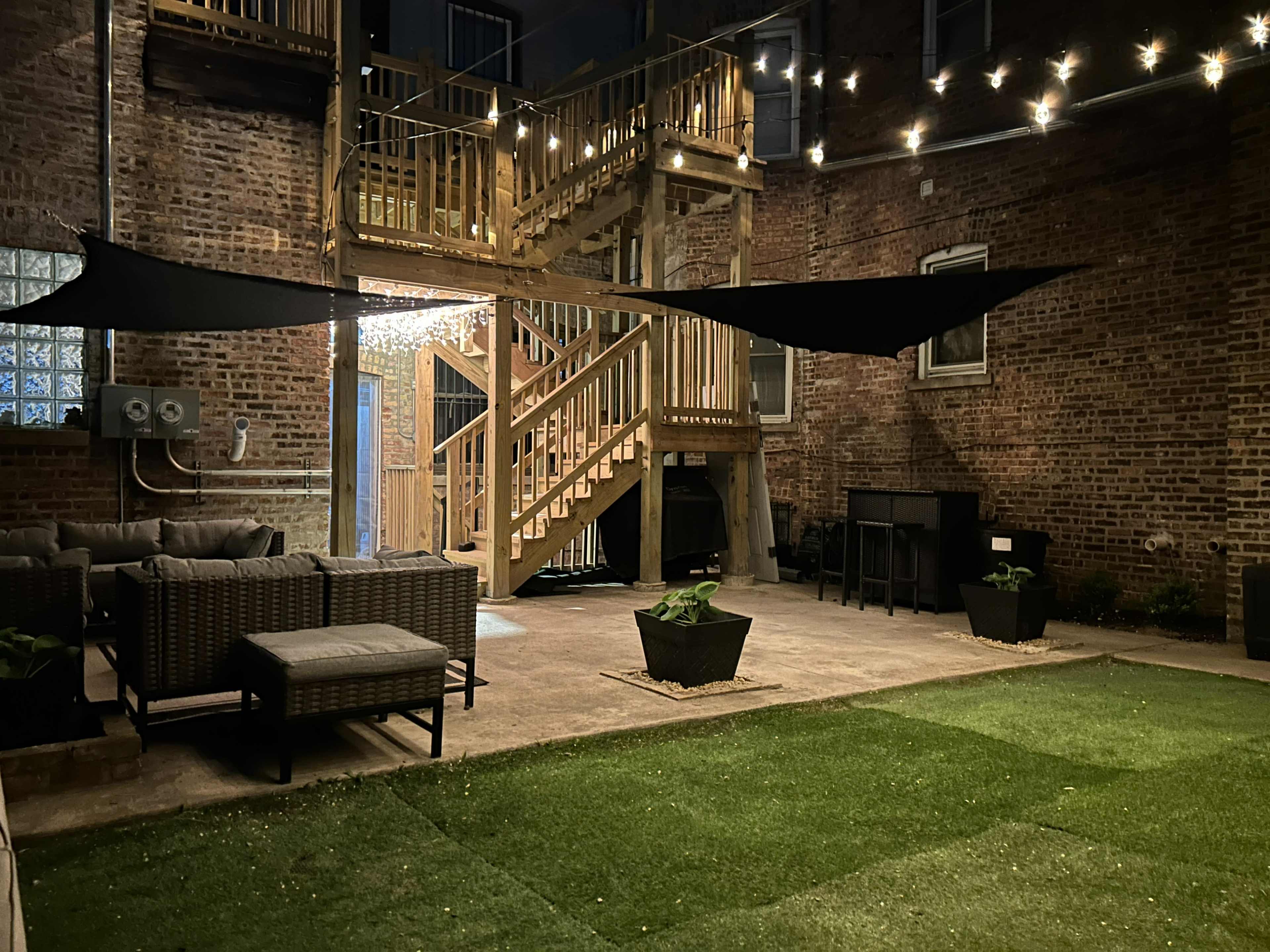 A well-lit courtyard featuring wooden stairs, lounge furniture, and potted plants under string lights.
