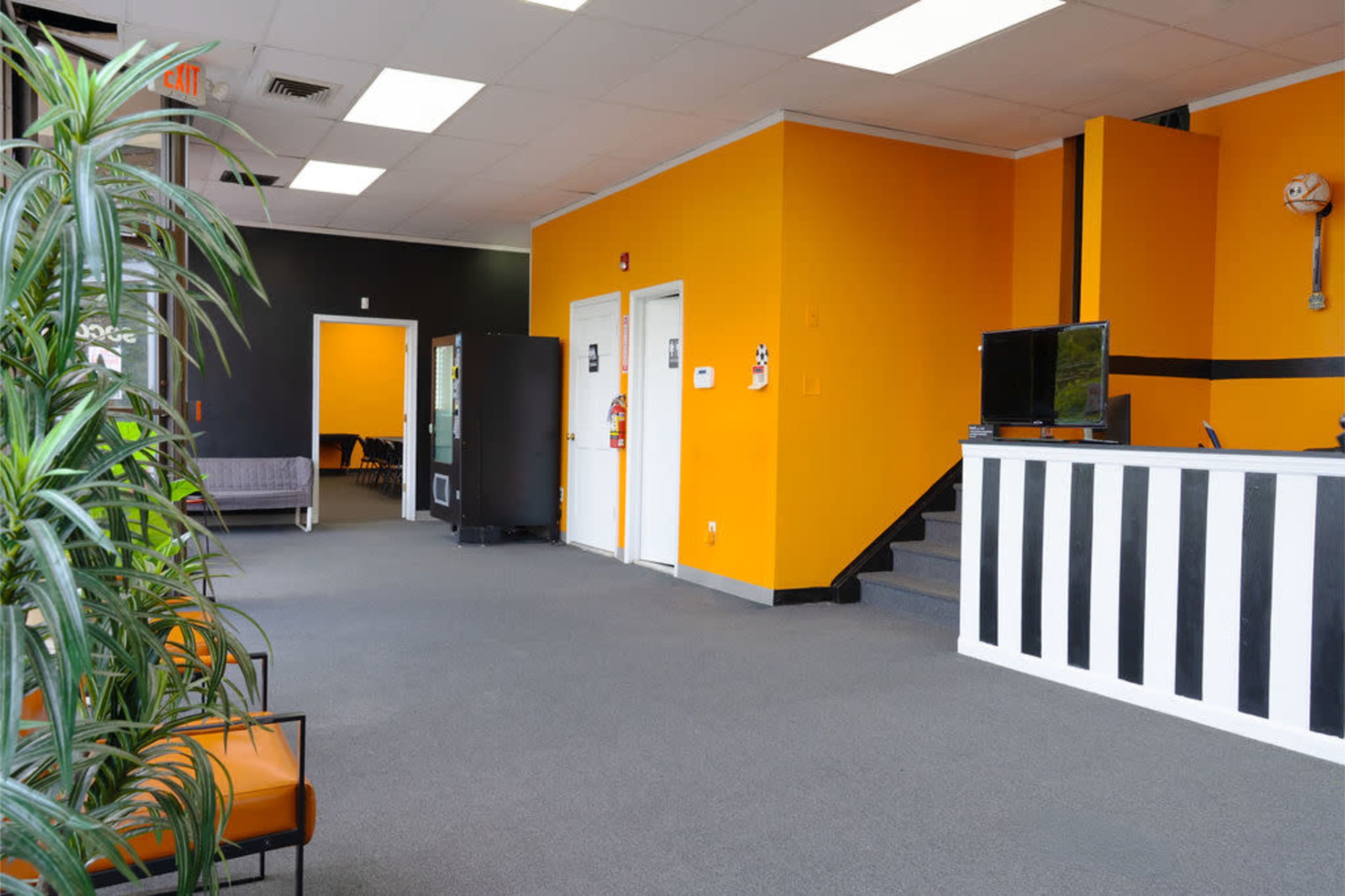 The image shows a modern interior space featuring a reception area with orange and black walls, a seating arrangement, a vending machine, and a staircase leading to an upper level.