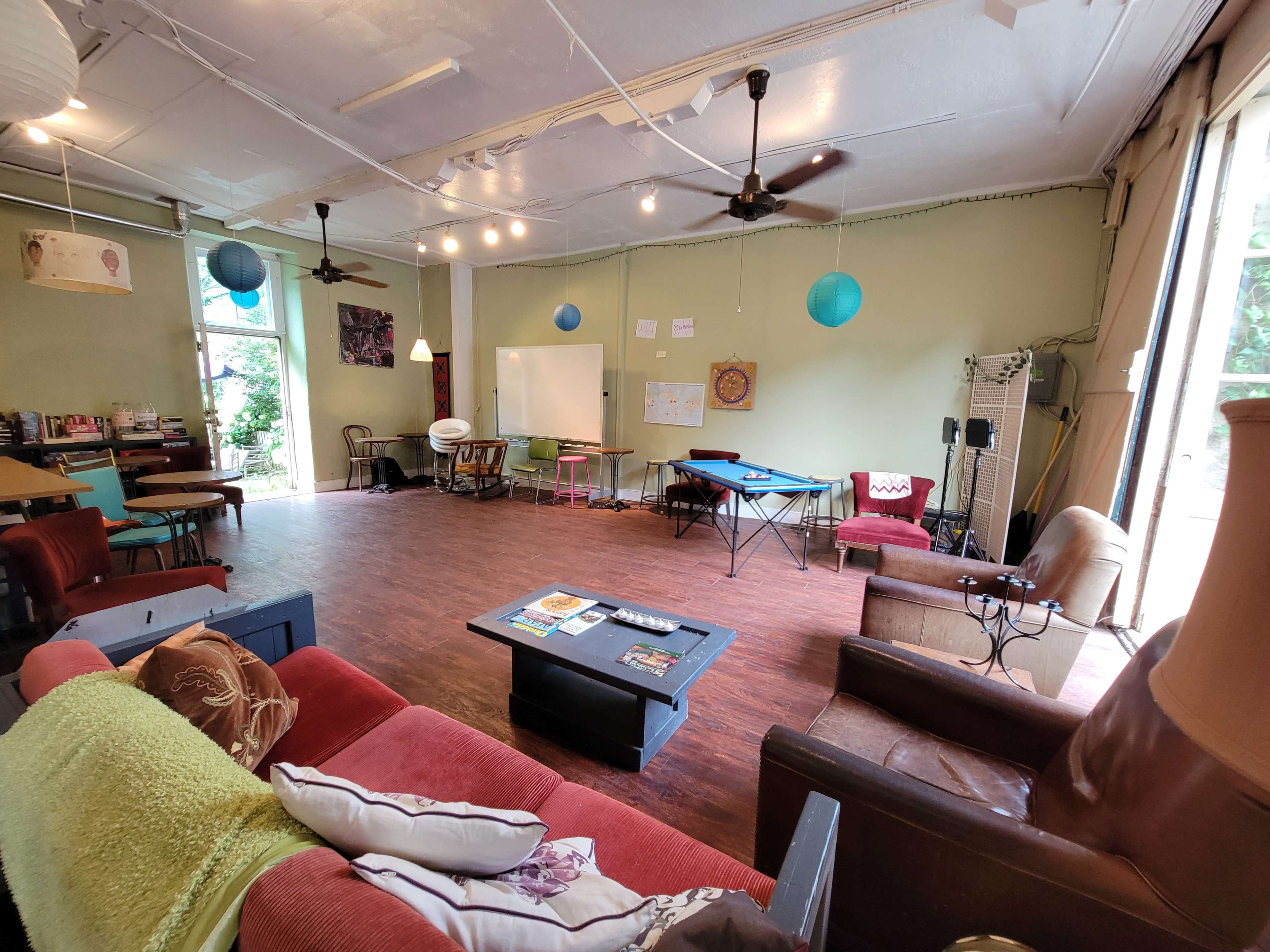 The image shows a spacious room with wooden floors and various seating areas, including couches and tables, illuminated by natural light from large windows.