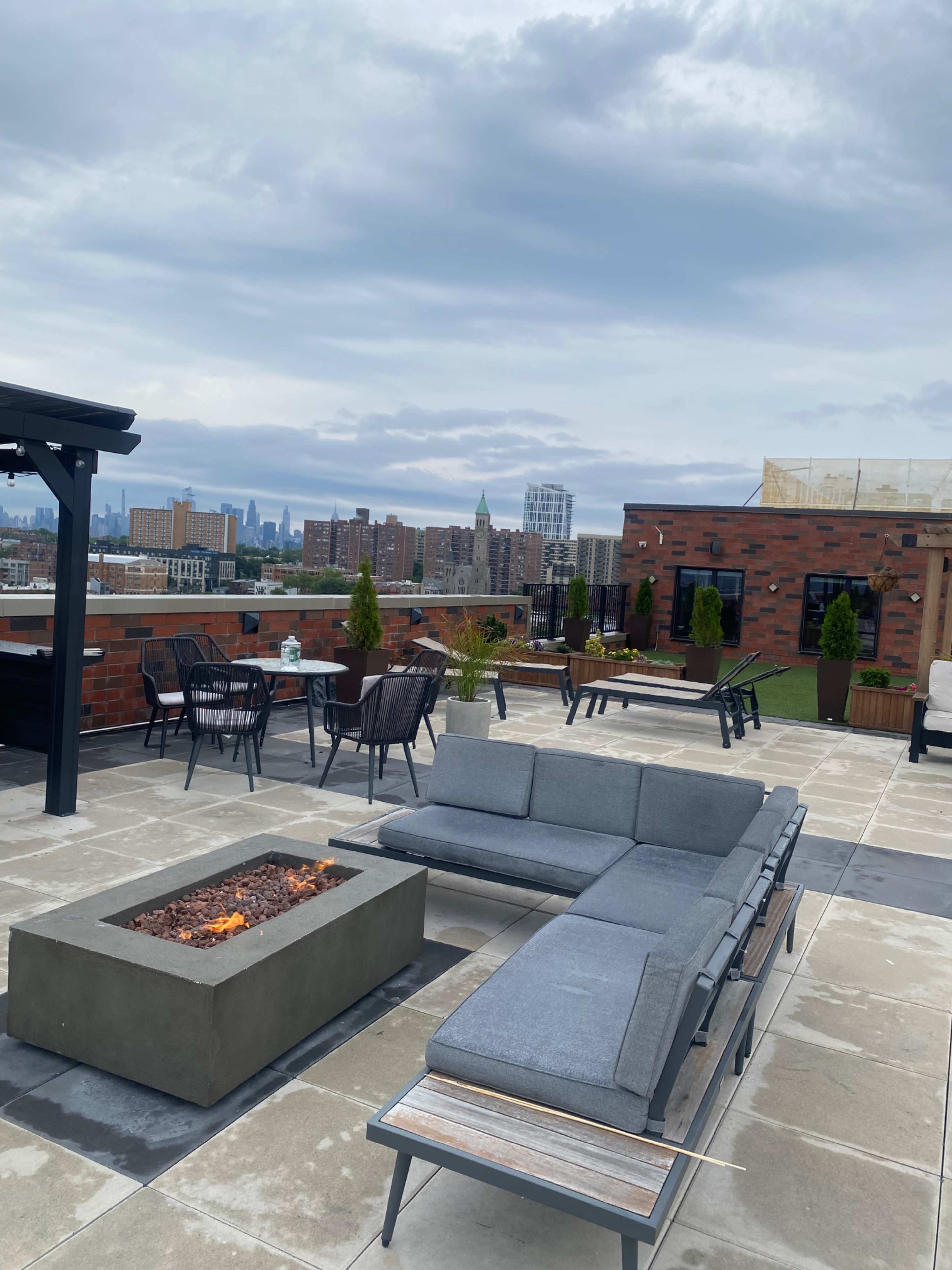 The image shows a rooftop patio featuring a gray sectional sofa, a fire pit, and various seating areas with views of a city skyline in the background.