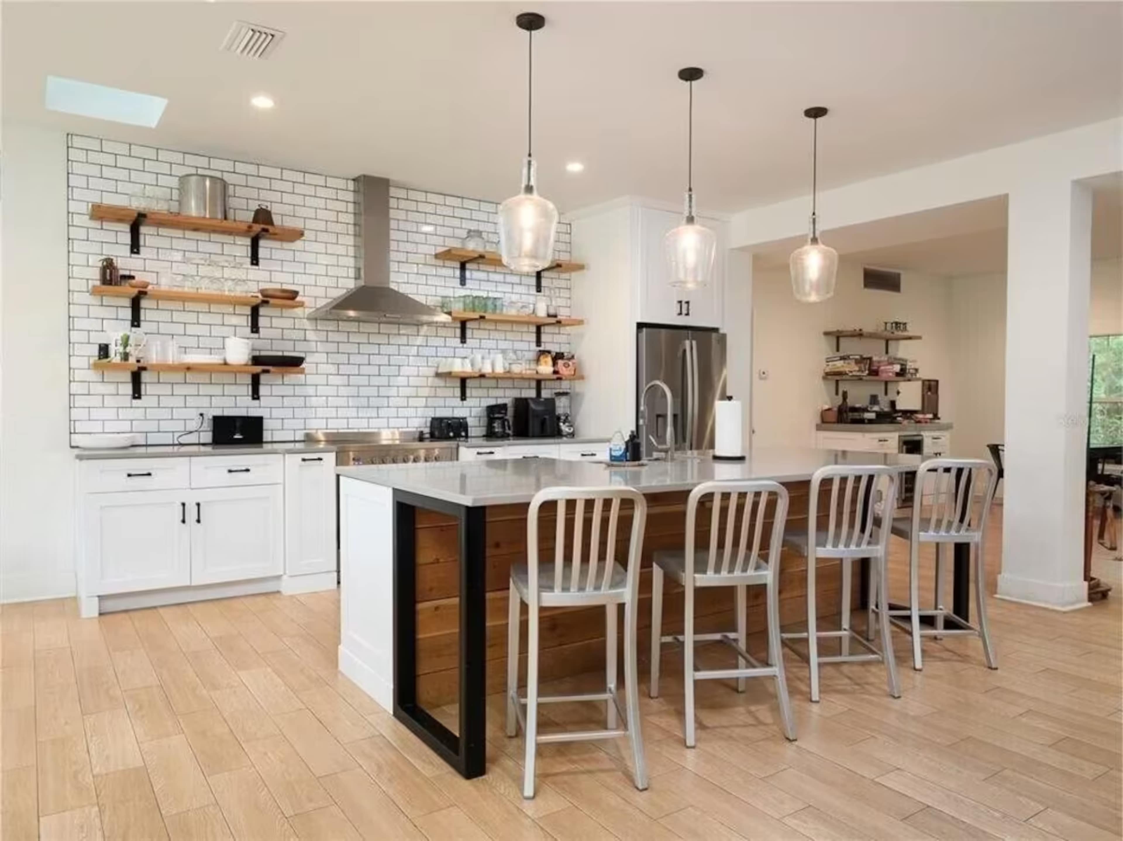 A modern kitchen with white cabinets, a gray island, wooden shelving, stainless steel appliances, and four bar stools.