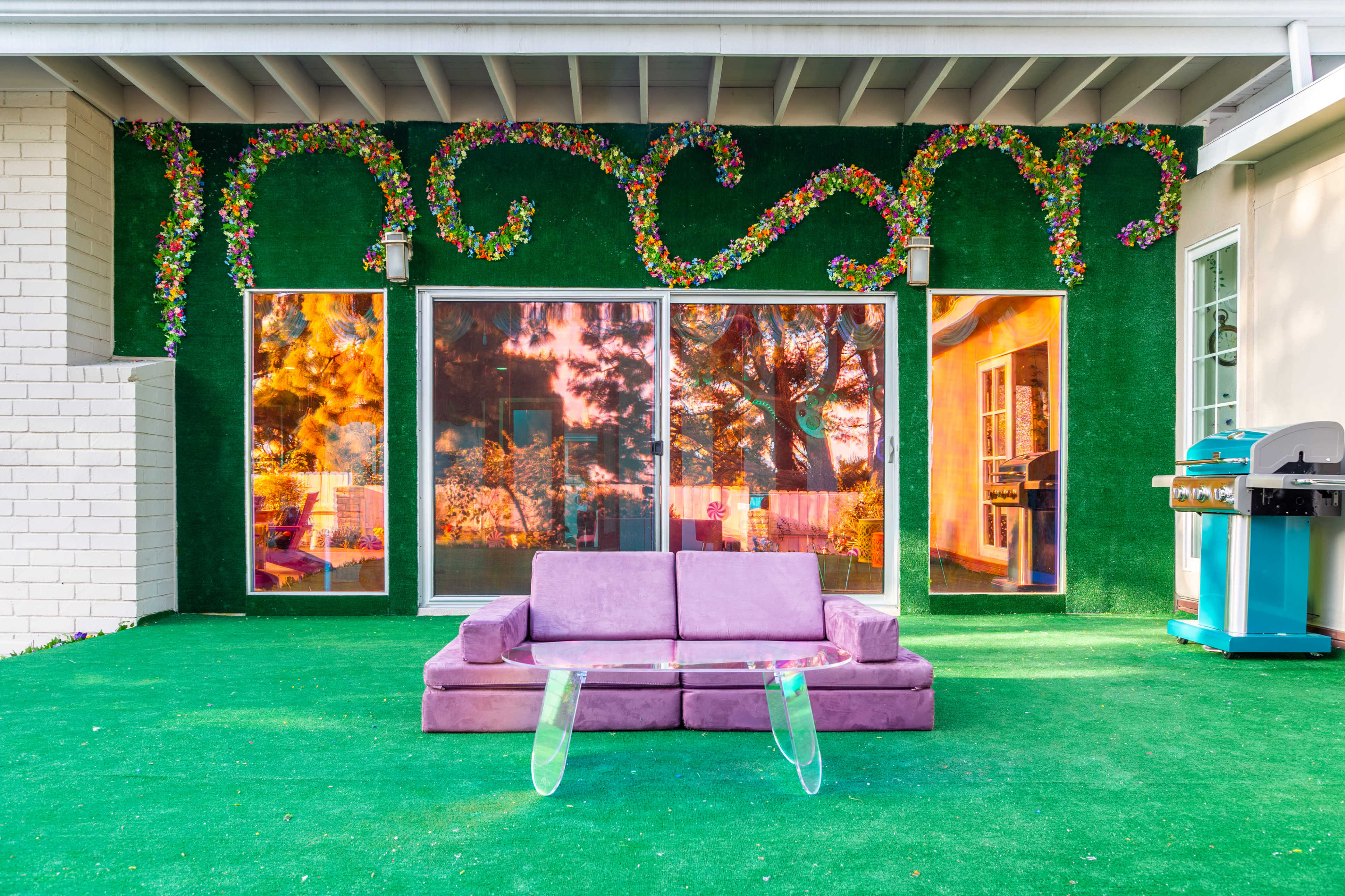 A purple couch sits on green turf in front of large windows adorned with floral decorations on a patio.