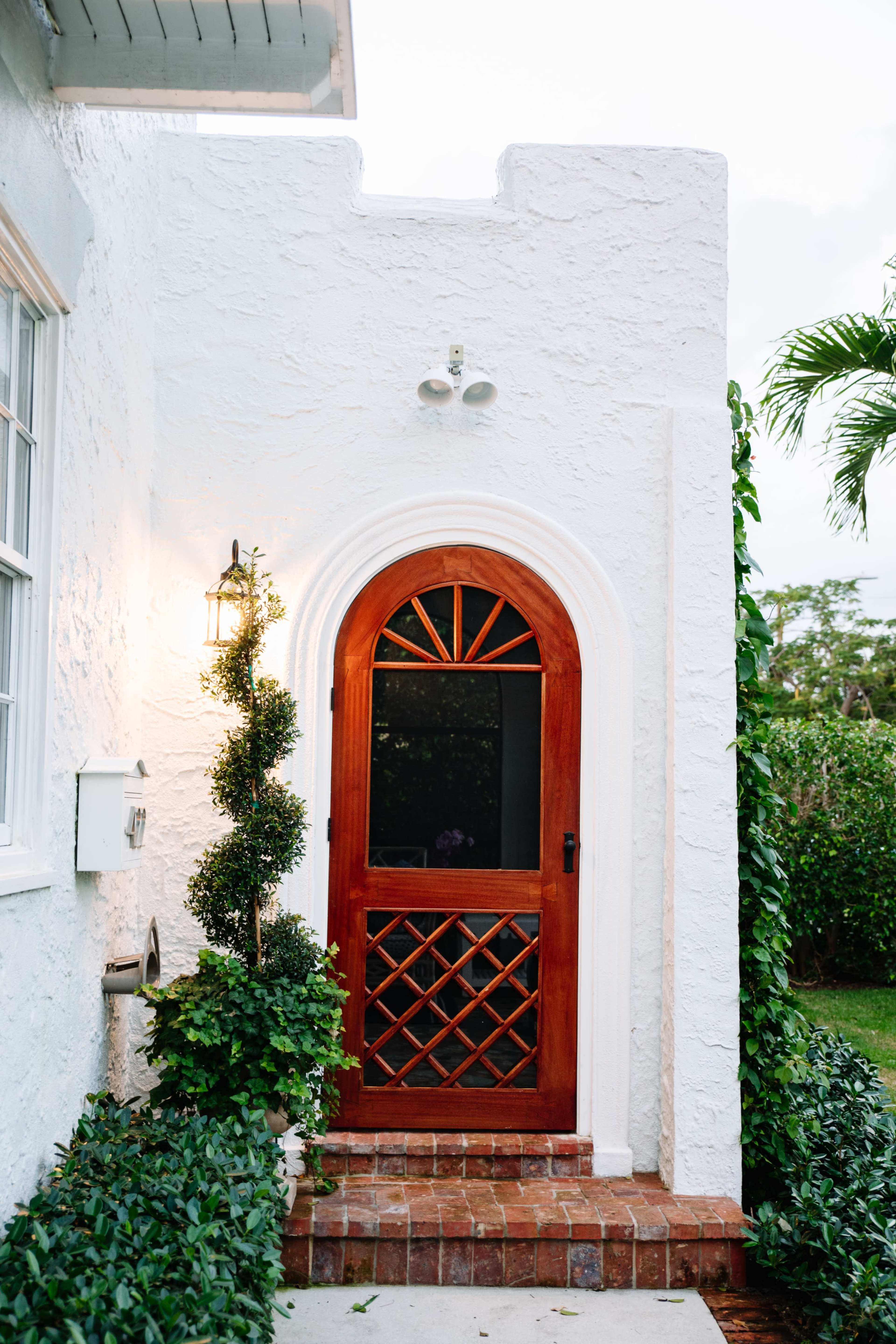 The image shows a white stucco house entrance featuring a wooden arched door, flanked by greenery and a wall-mounted lantern.
