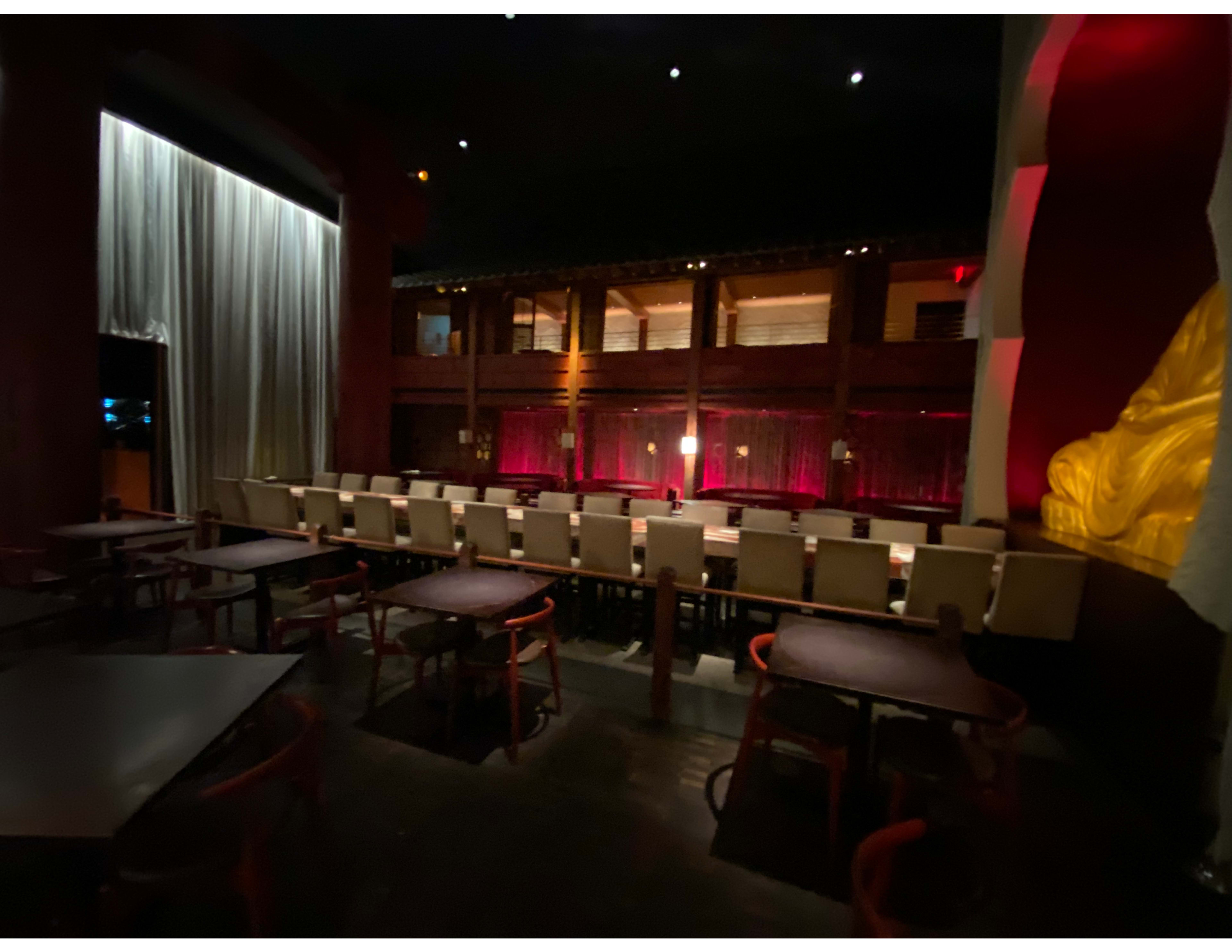 A dimly lit restaurant interior featuring a long table with chairs, a stage area in the background, and decorative curtains on the side.