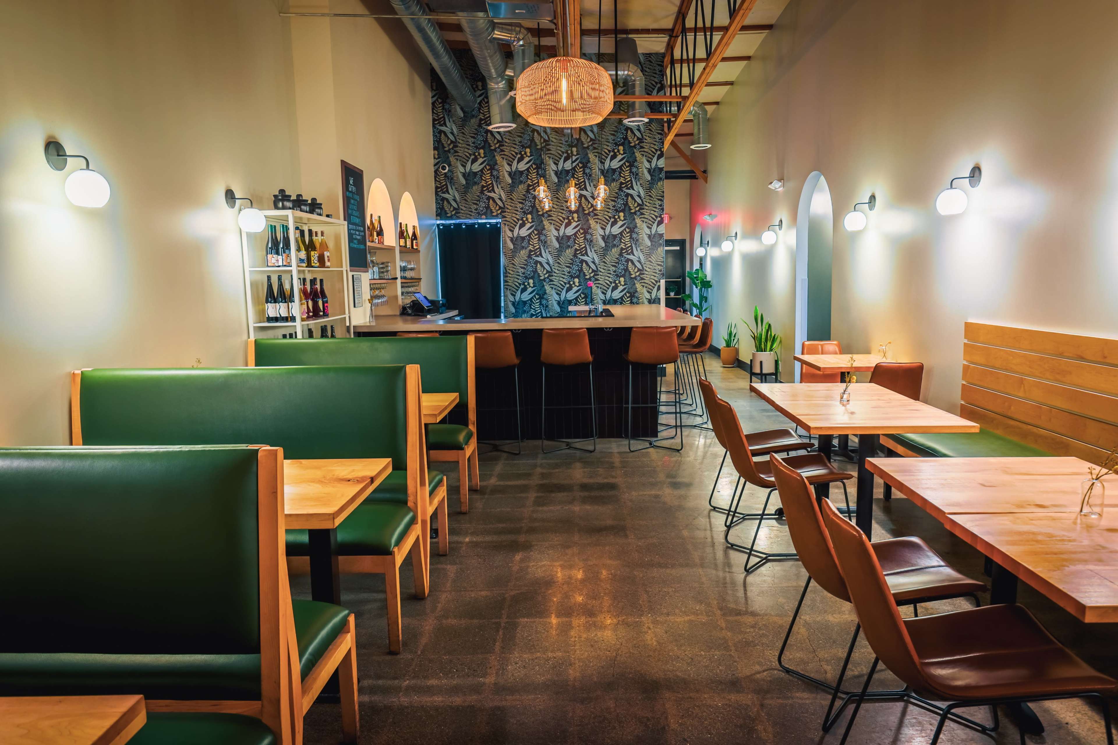 The image shows a modern restaurant interior with green leather seating, wooden tables, and a bar area featuring a patterned wallpaper and pendant lighting.