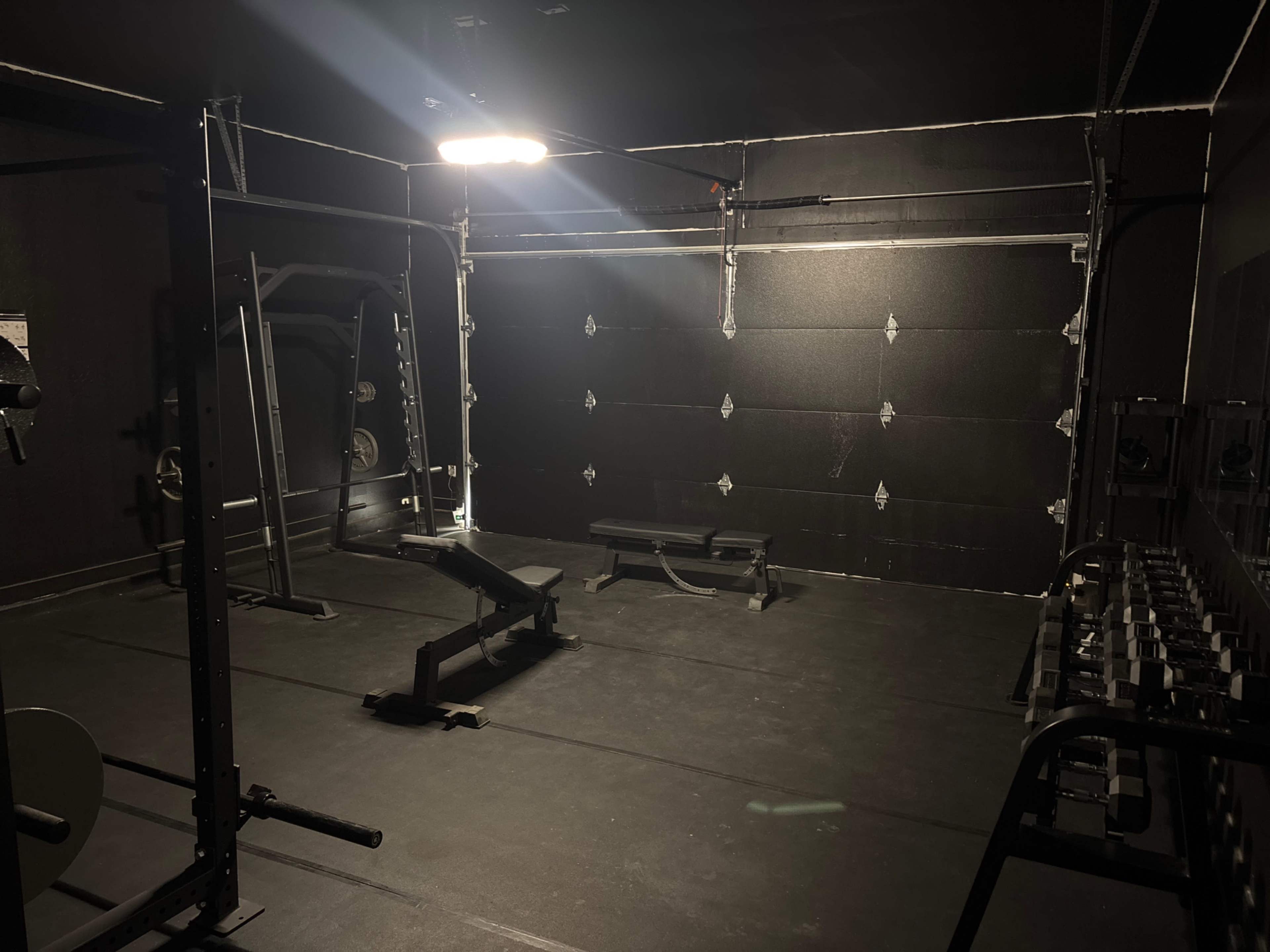 The image shows a dimly lit gym space featuring exercise equipment, including a bench, weights, and a squat rack against a dark backdrop.