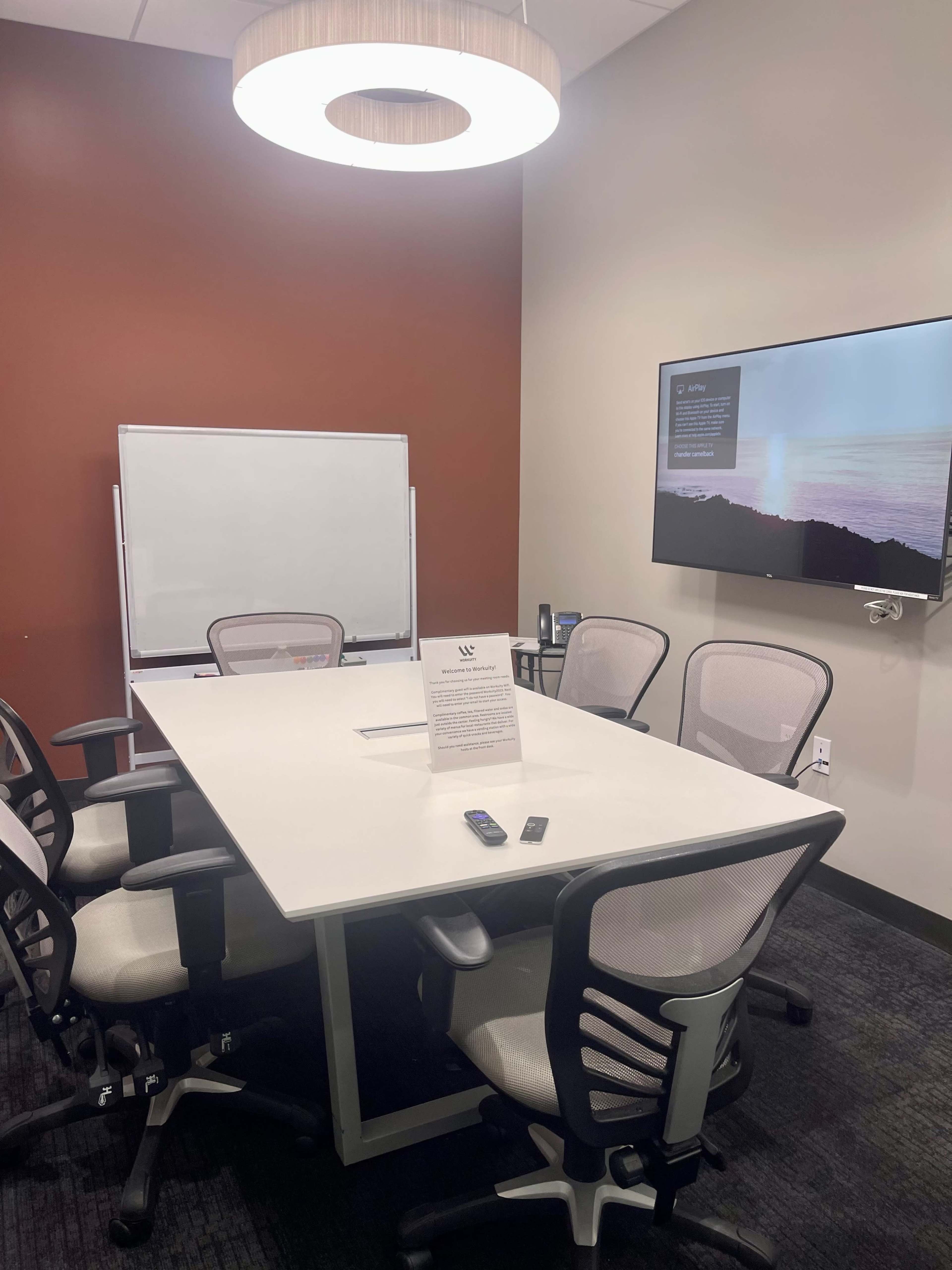 A small meeting room with a rectangular table, six chairs, a whiteboard, and a wall-mounted television.