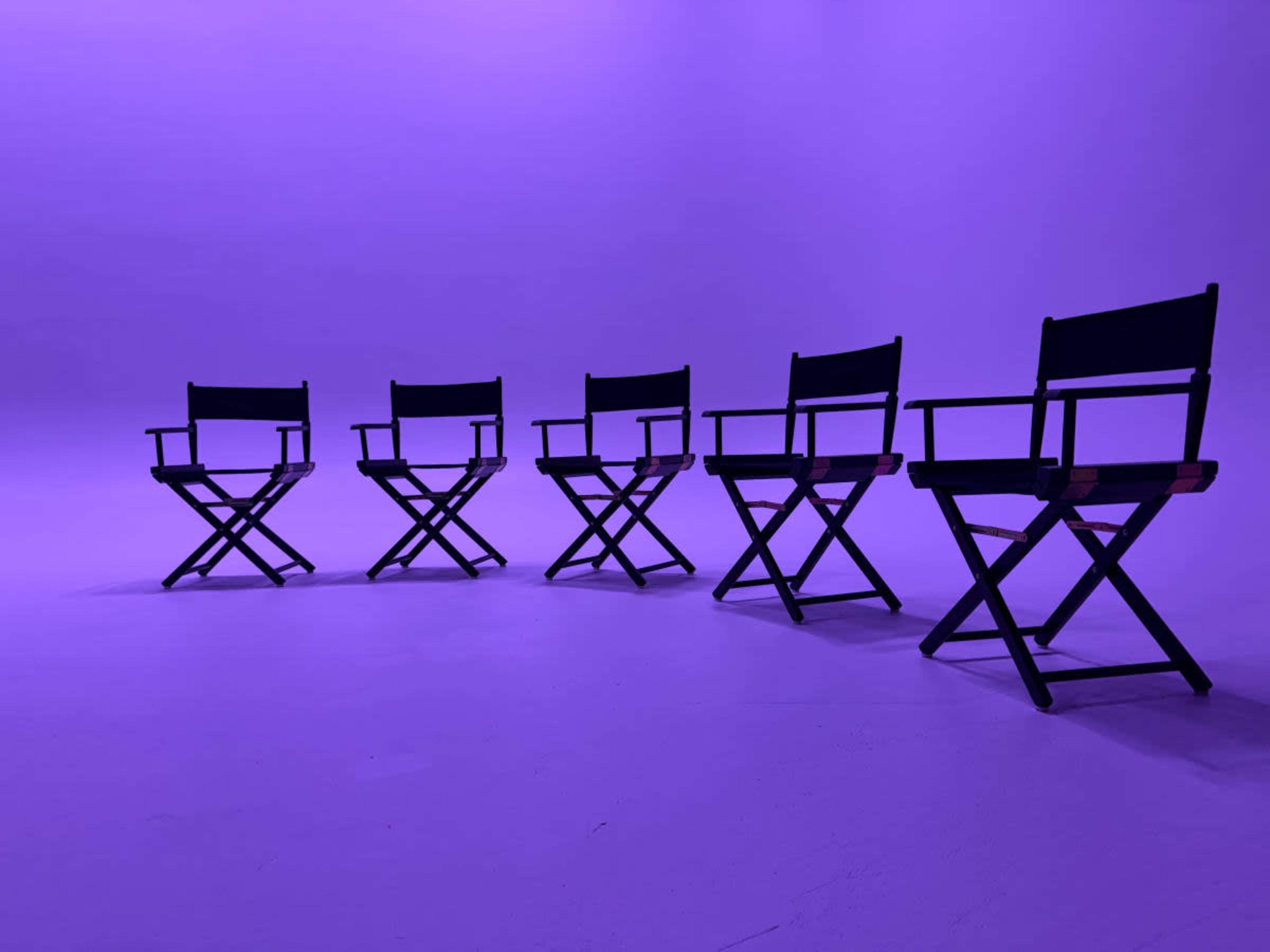 Five black director's chairs are arranged in a row against a purple backdrop.