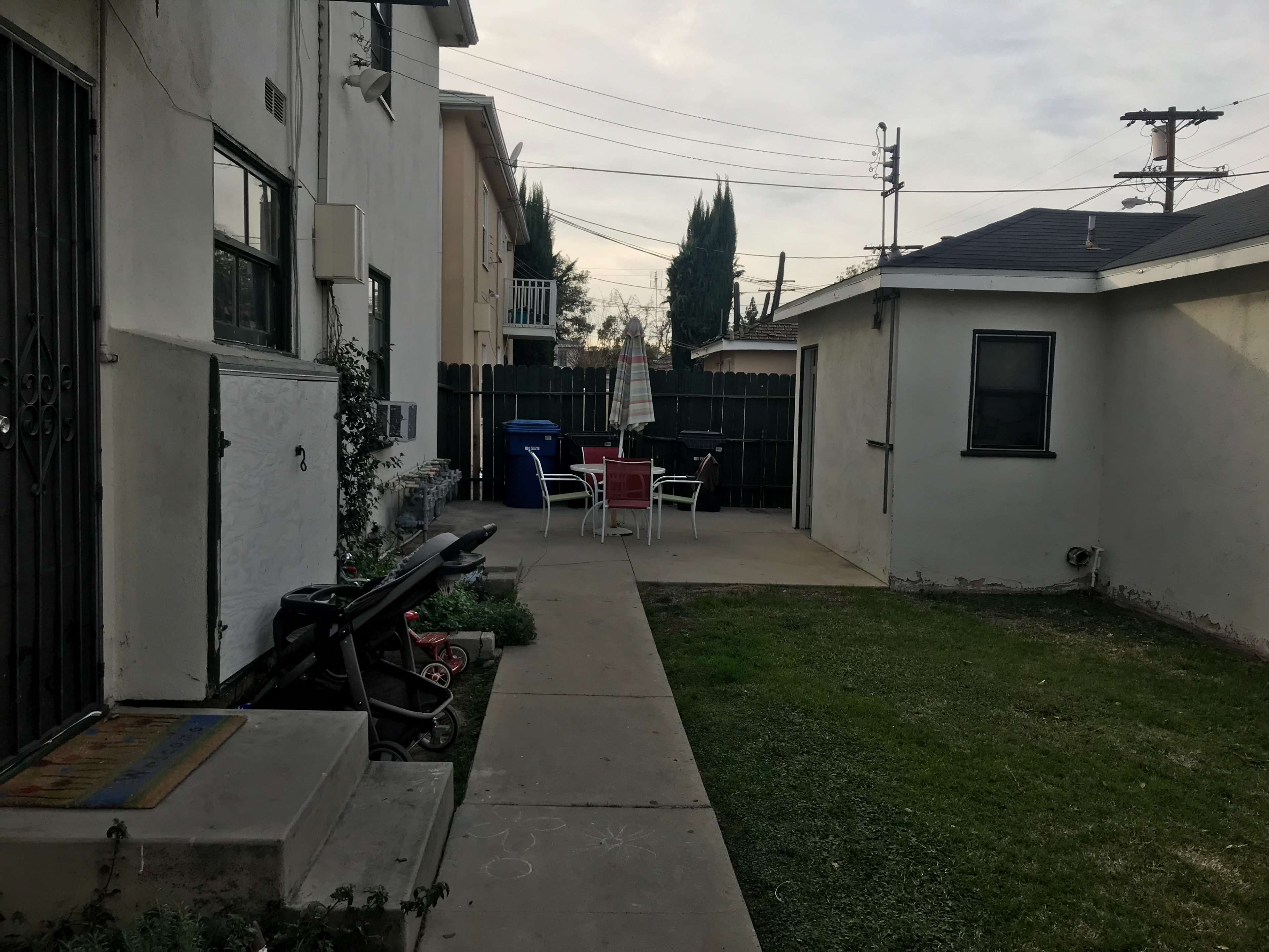 The image shows a narrow pathway beside a building, leading to a patio area with a table and chairs, and a grassy section on one side.