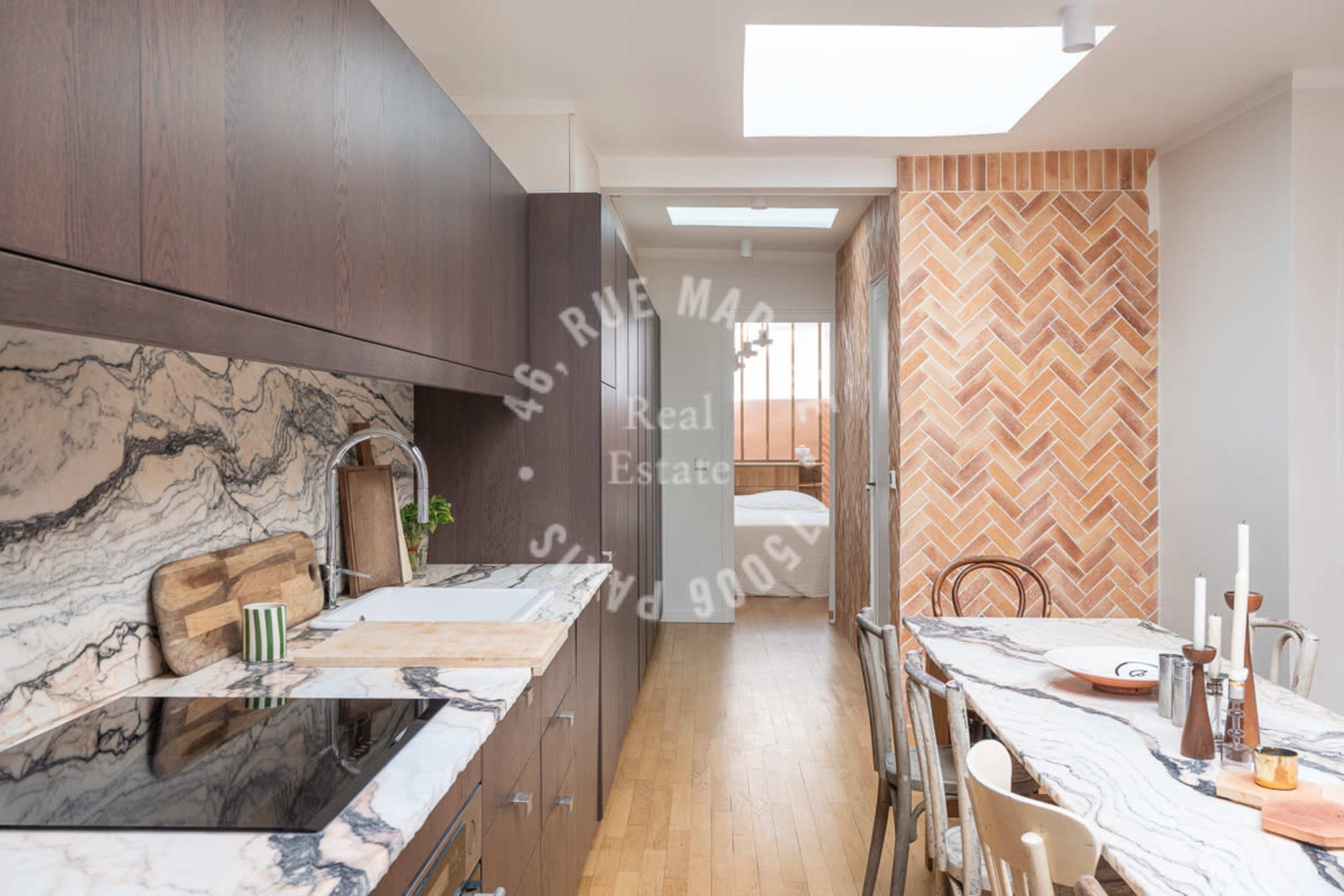 The image shows a modern kitchen with a marble countertop, wooden cabinetry, and a dining area featuring a table set with candles.