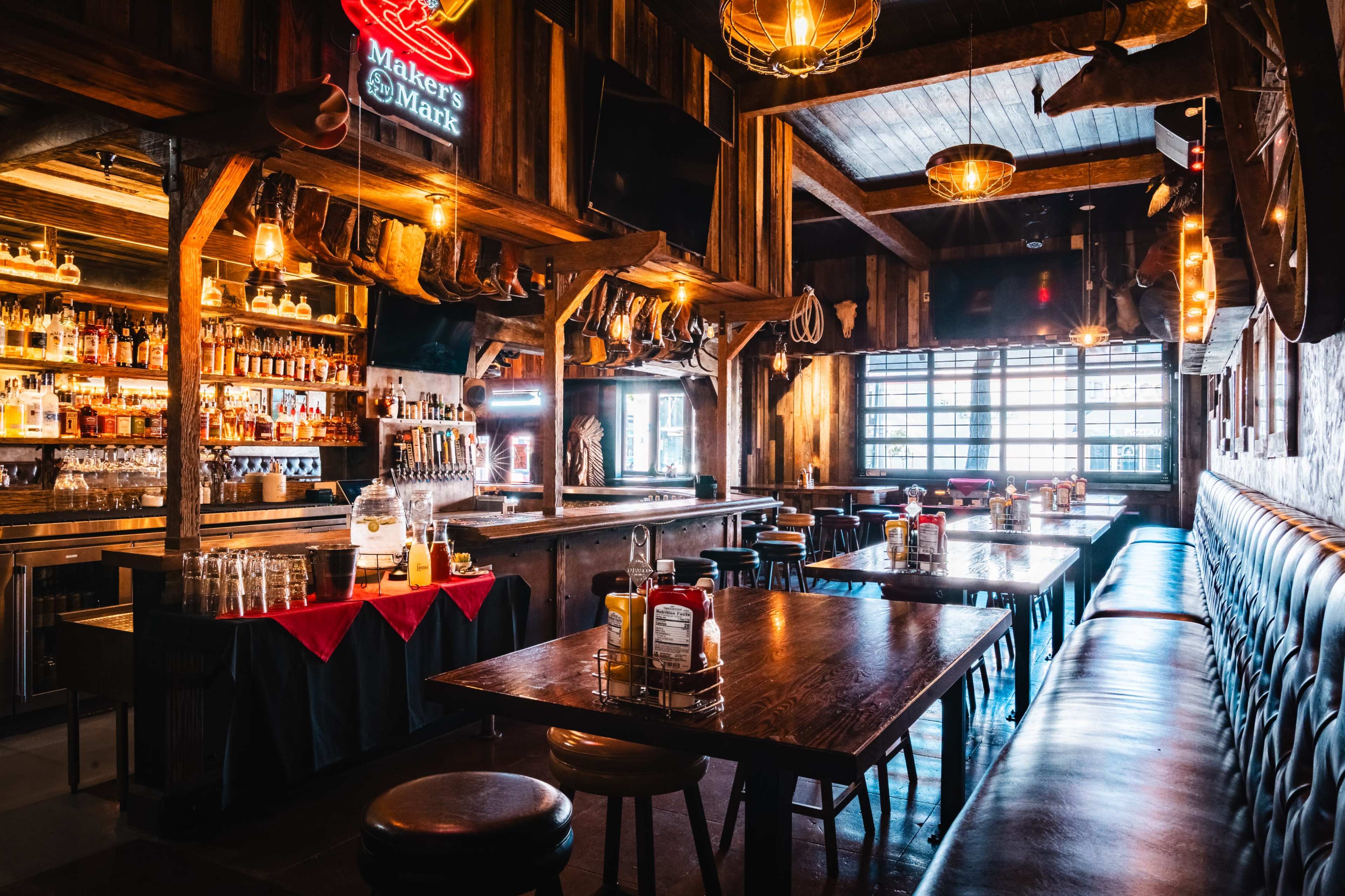 The image shows a rustic bar interior with wooden beams, a long wooden table, and a well-stocked bar featuring various bottles and dim lighting.