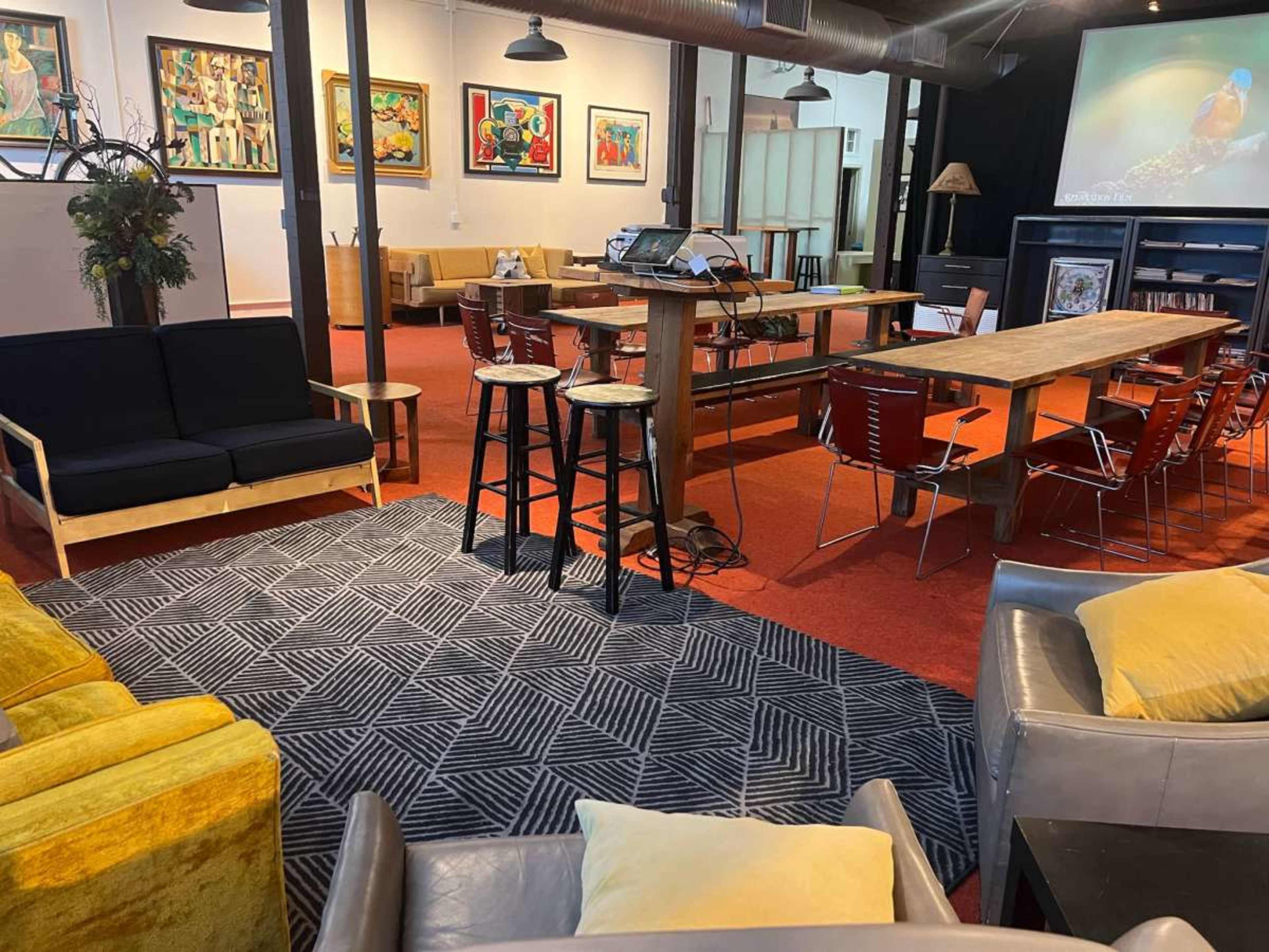 The image shows an interior space with various seating arrangements, including a black couch, a long wooden table, and several metal chairs, all set against a backdrop of colorful artwork on the walls.