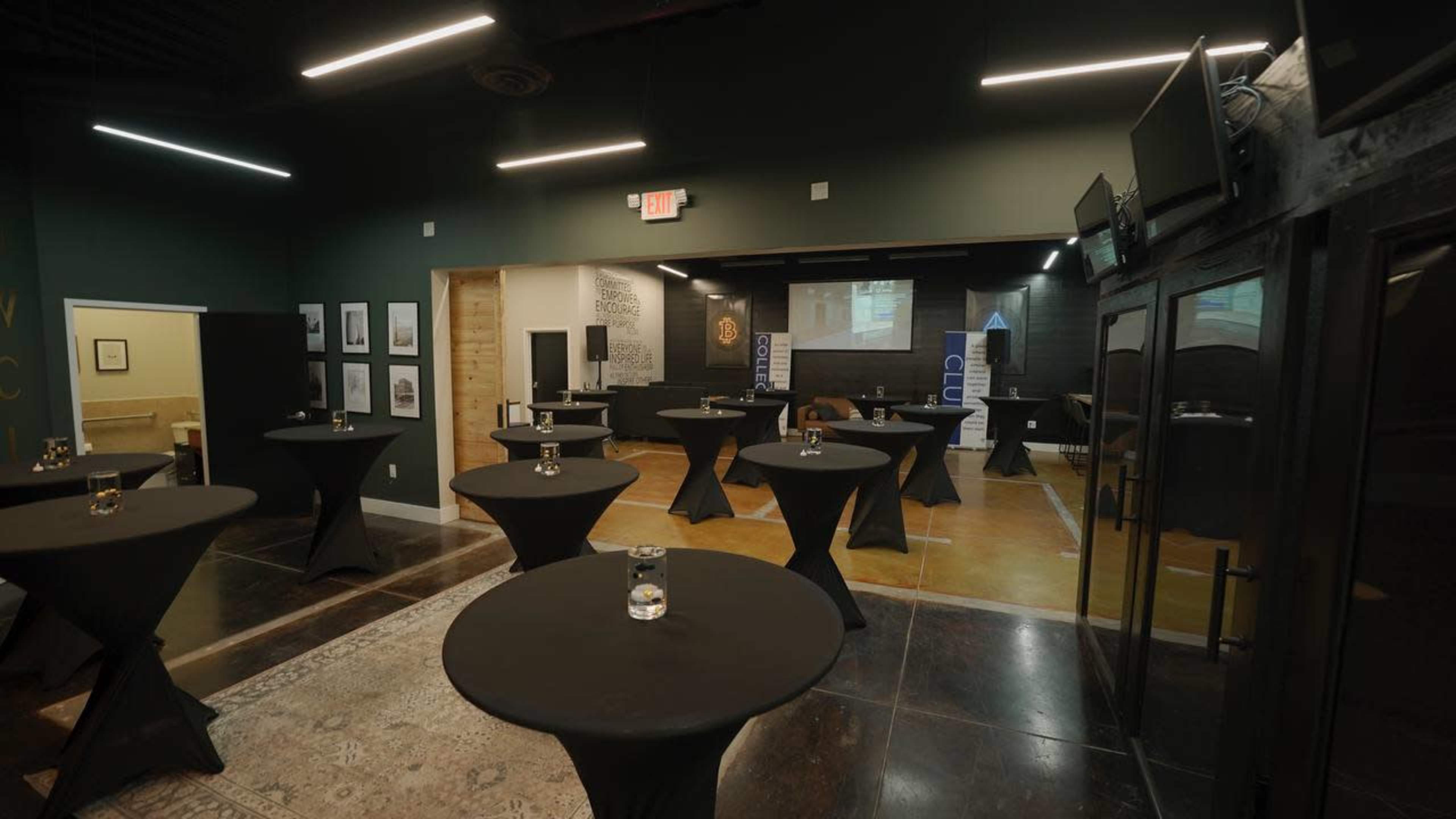 A modern event space with several round tables covered in black linens, arranged around a central area, and a projection screen at the front.