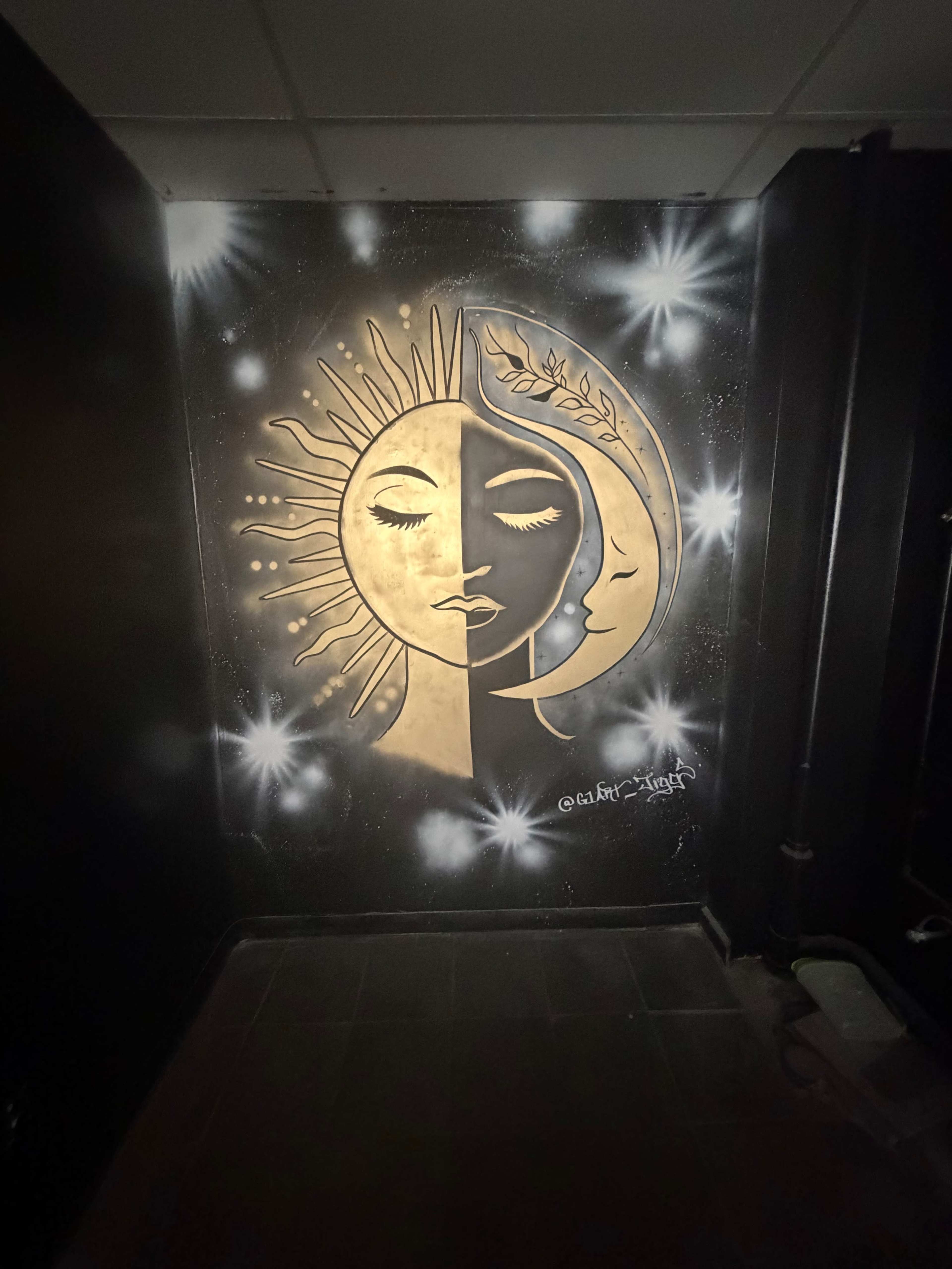 The image shows a mural of a half sun and half moon, intricately designed in gold against a black wall, surrounded by star-like patterns.