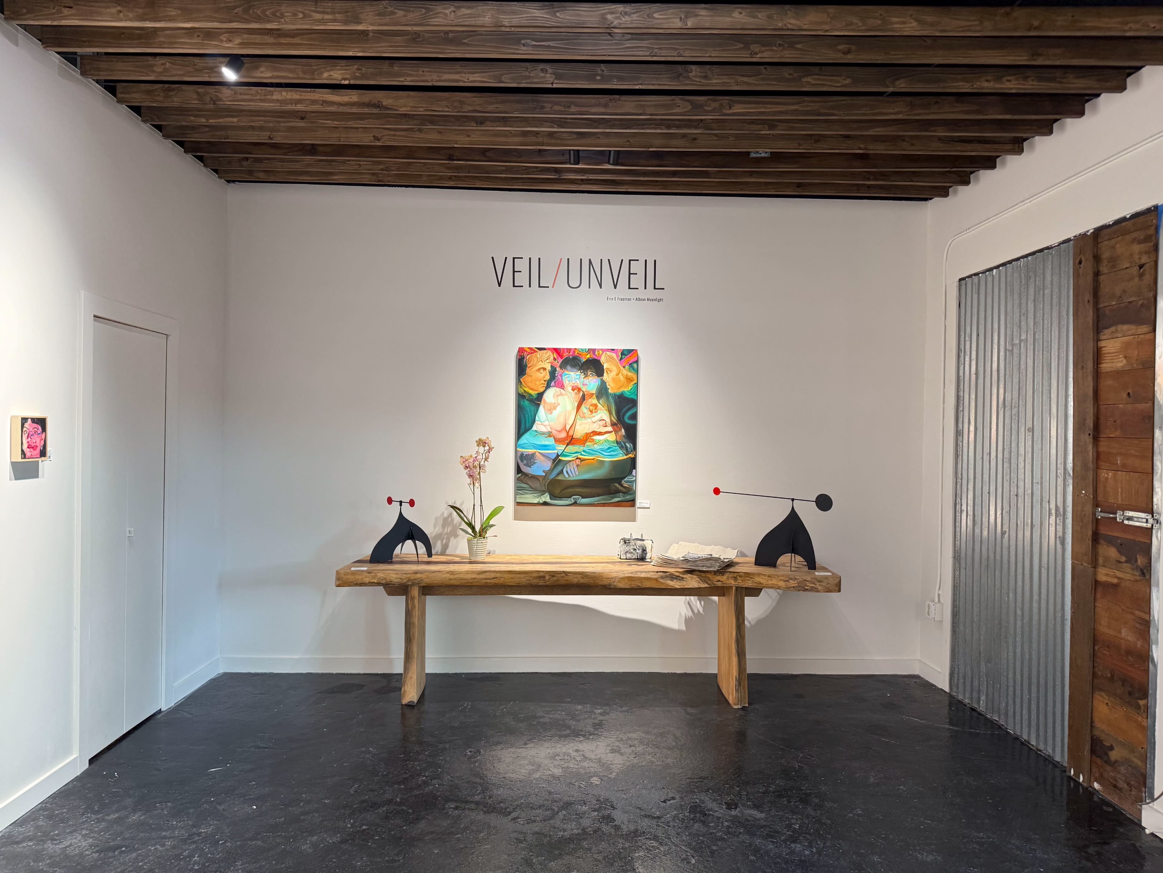 The image shows an interior gallery space featuring a large painting of a figure on the wall, a wooden table with decorative items, and two sculptural elements on either side of the table.