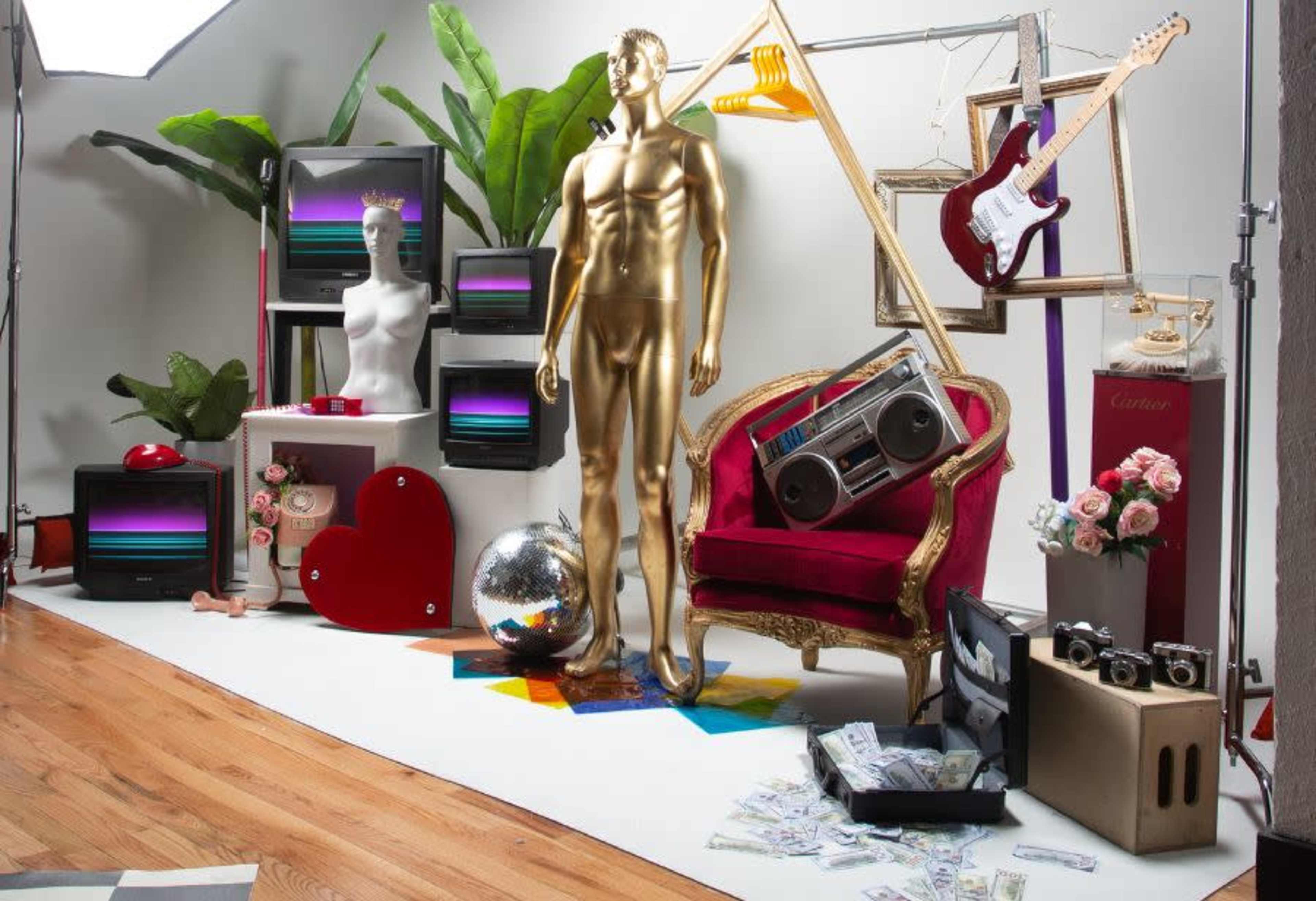 The scene features a gold mannequin standing among vintage televisions, a disco ball, a red heart sculpture, a retro boombox, a plush chair, and various decorative elements.