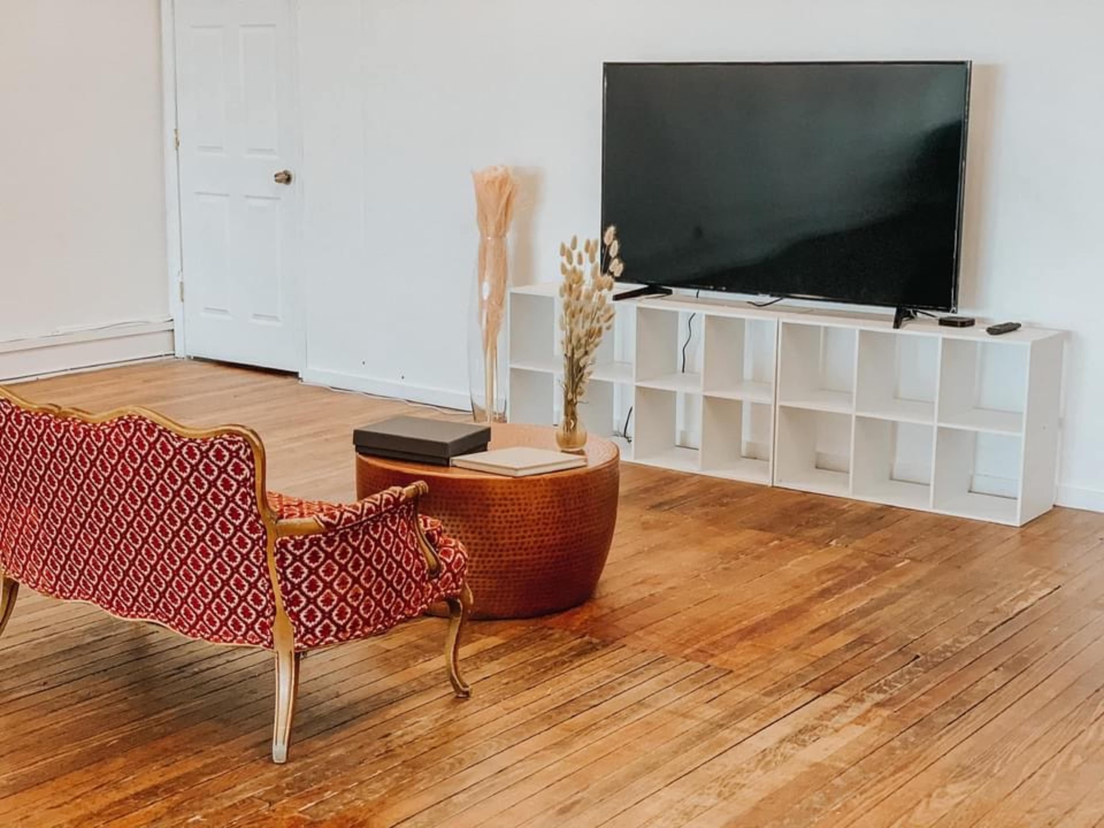 A red upholstered couch faces a large flat-screen TV on a white media console in a minimally furnished room with wooden floors.