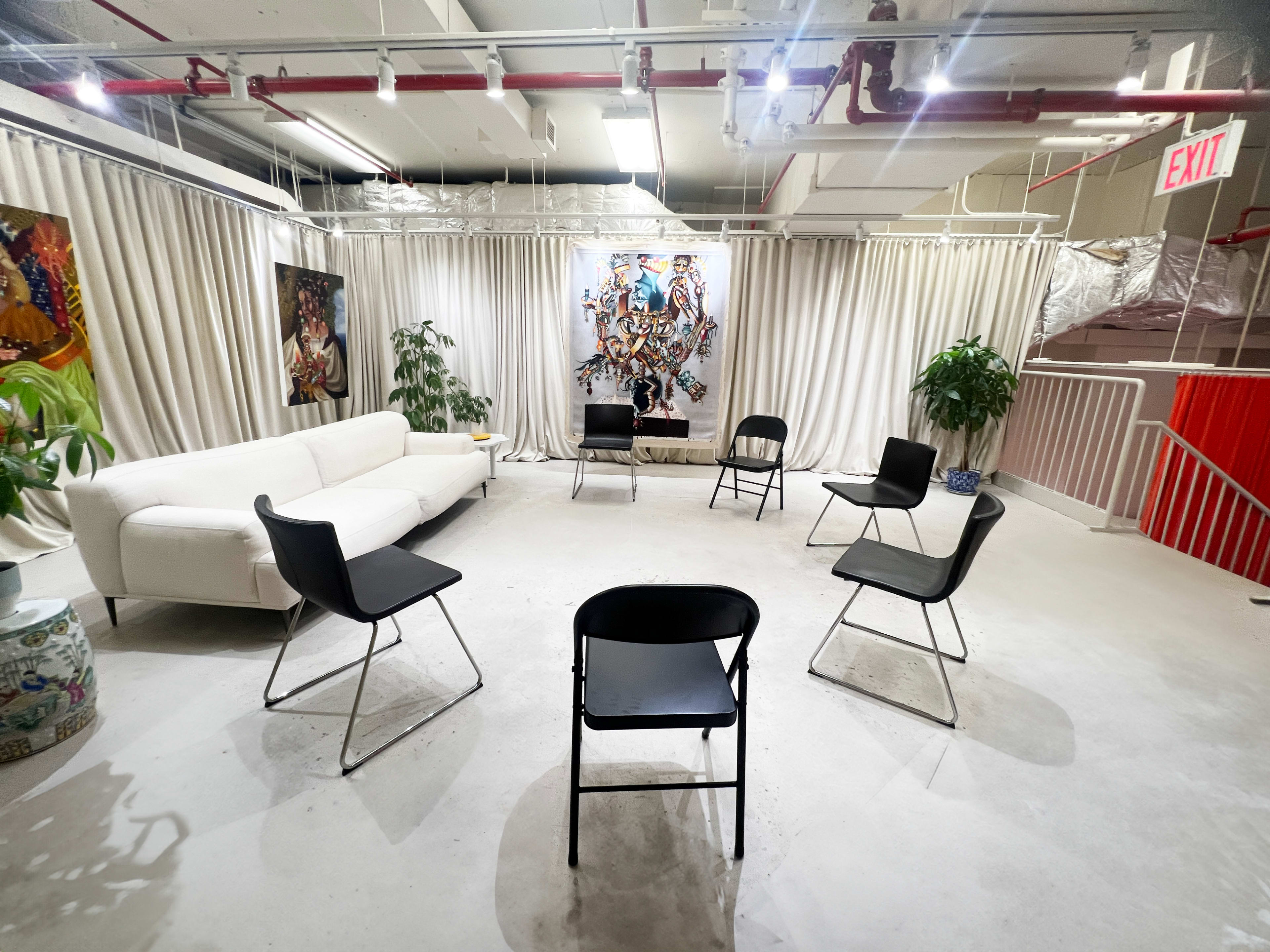 A minimalist meeting room with a white sofa and a circular arrangement of black chairs, surrounded by curtains and adorned with colorful artwork on the walls.
