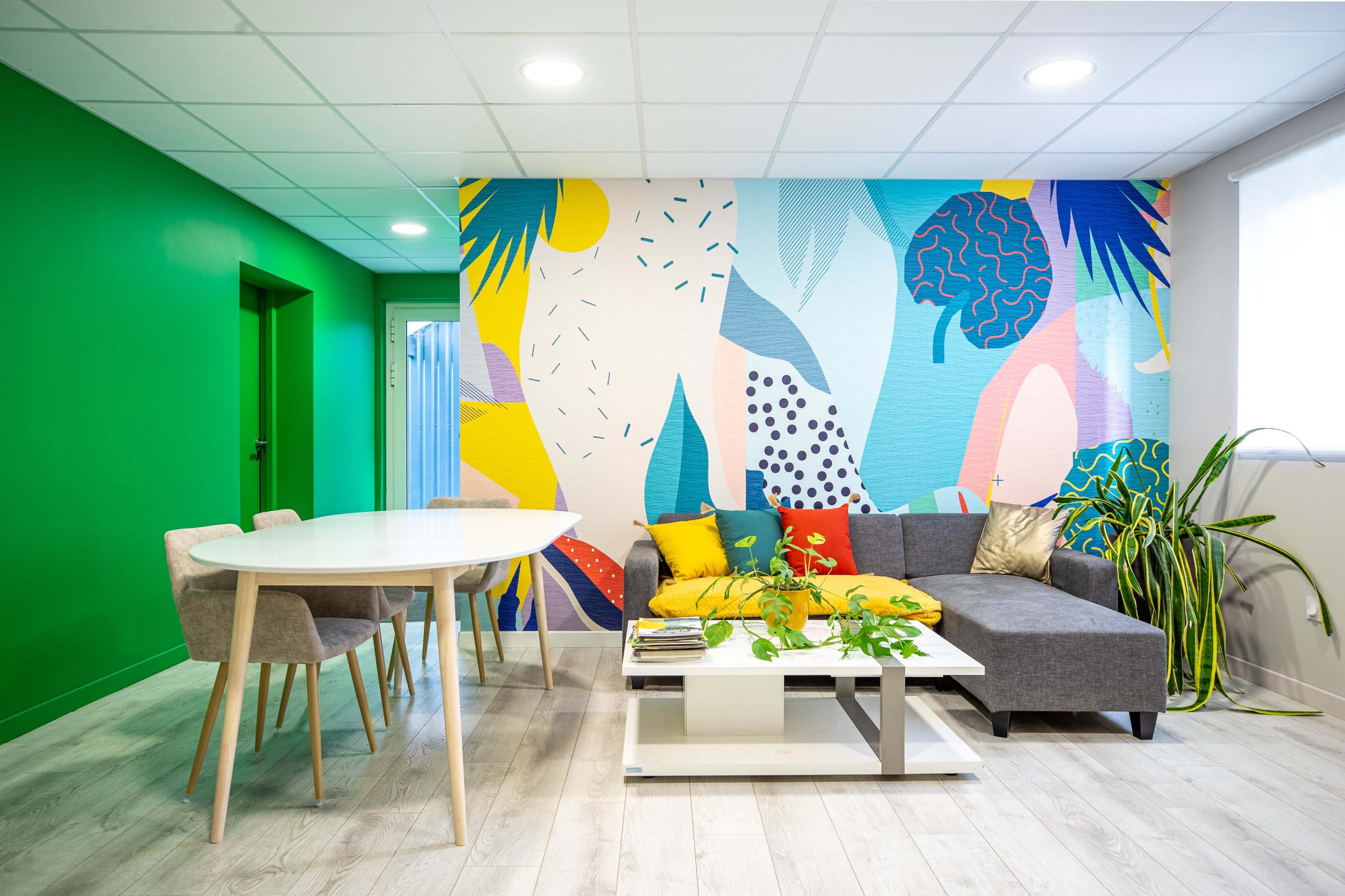 A modern living space features a colorful mural, a gray sofa with decorative pillows, a white coffee table, and a round dining table surrounded by chairs.