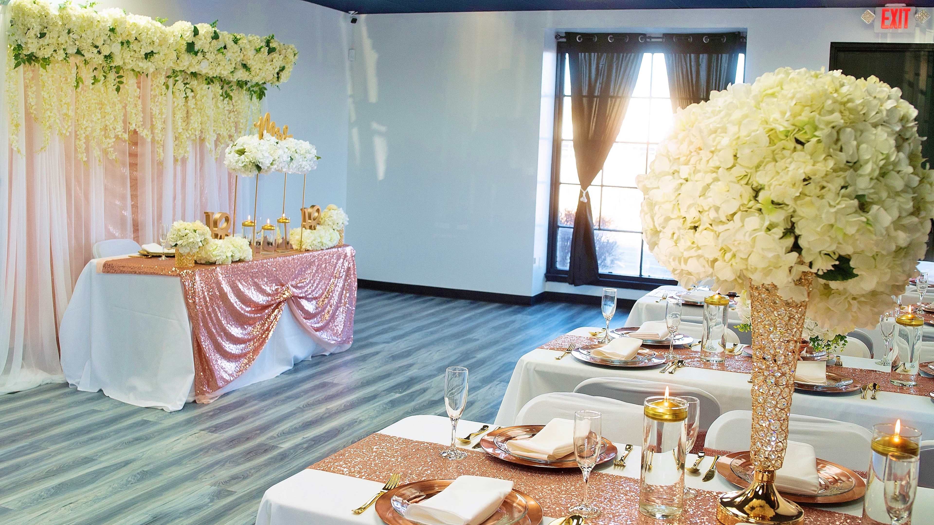 An elegantly decorated event space featuring tables set with white linens, gold accents, and large floral centerpieces, alongside a backdrop of cascading flowers and a shiny tablecloth.
