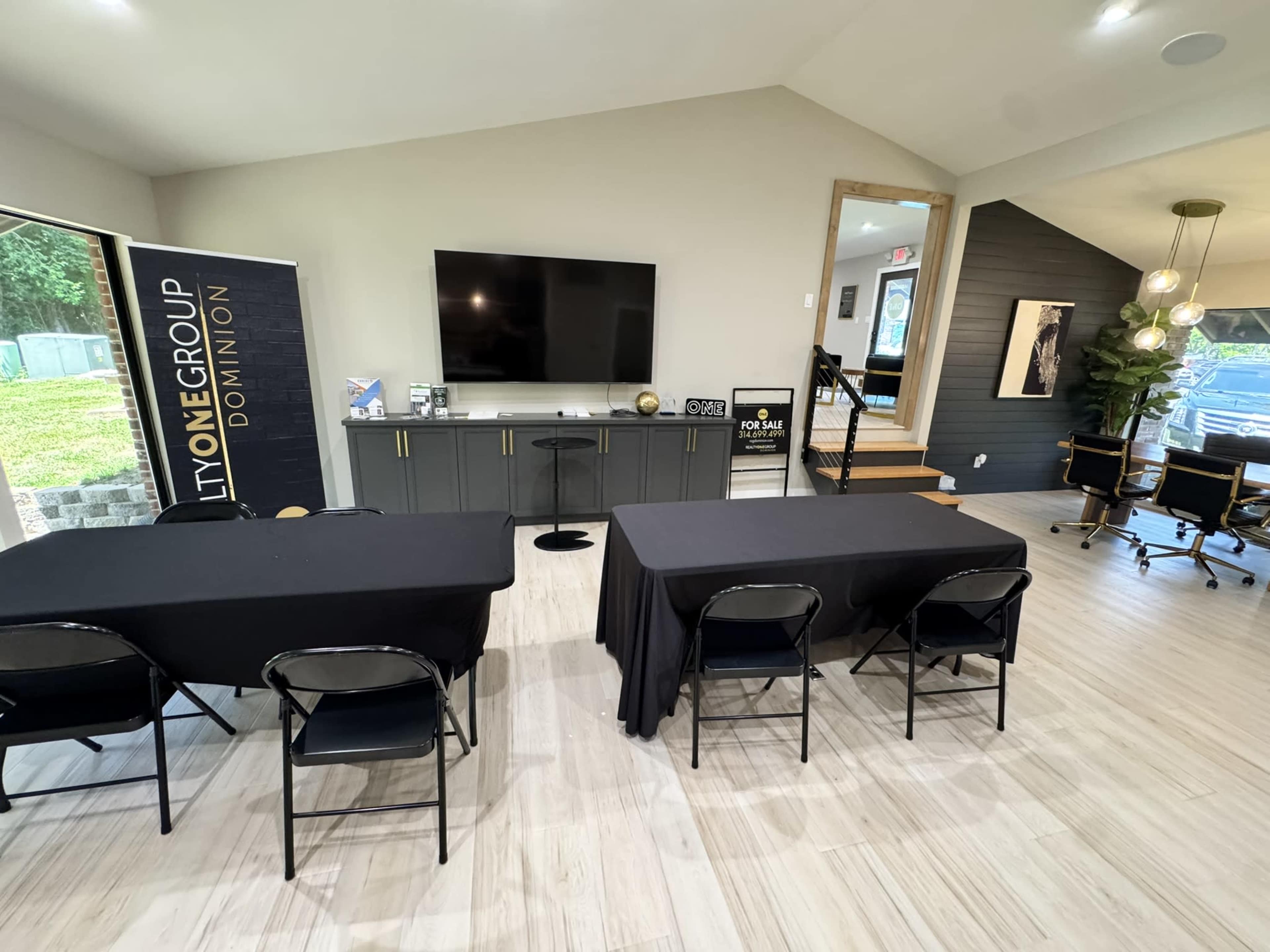 The image shows a modern meeting room with two tables covered in black cloth, a large television on the wall, and a staircase leading to an office area.