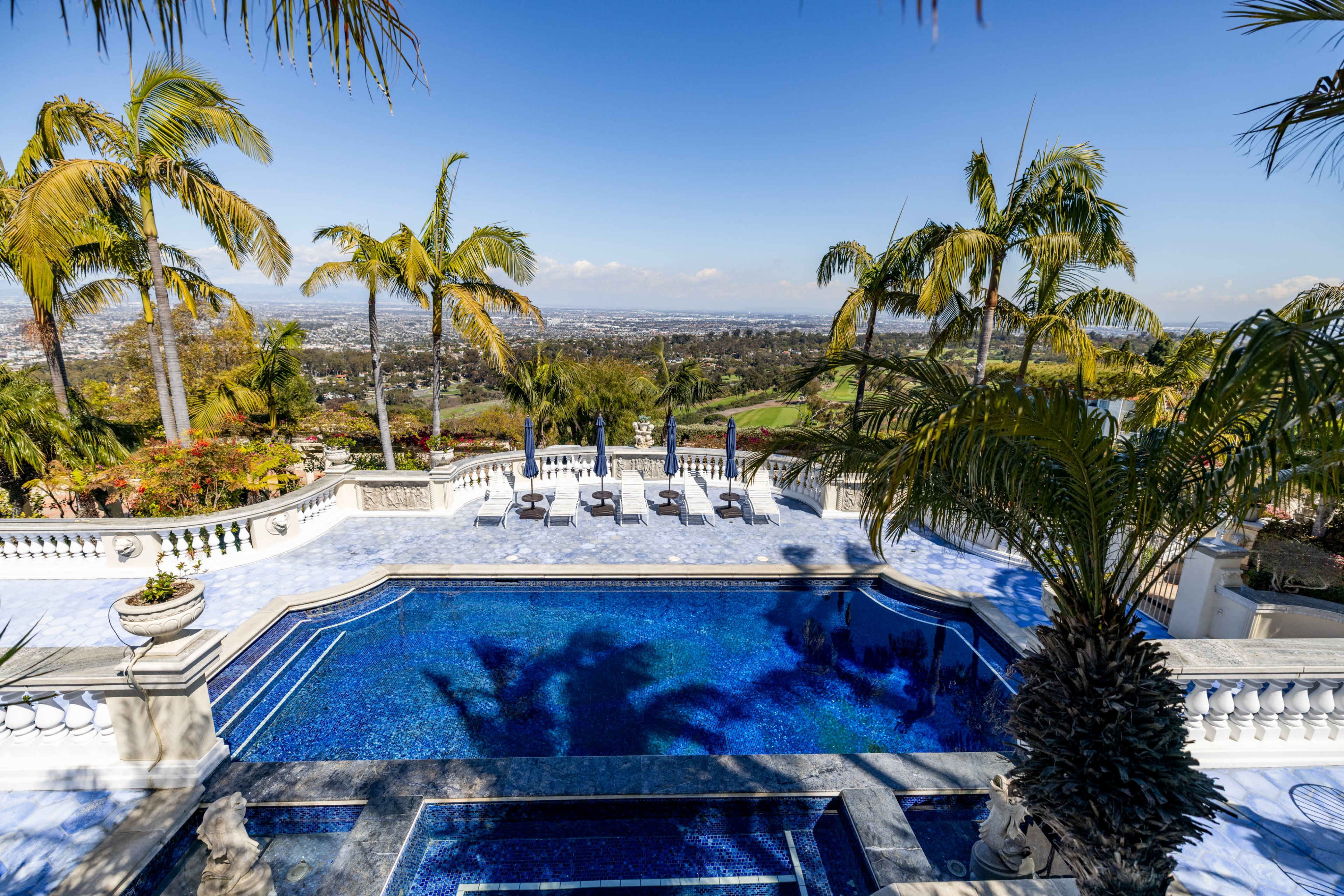 The image shows a luxurious outdoor area featuring a blue-tiled swimming pool, surrounded by palm trees and a scenic view of a hillside landscape.