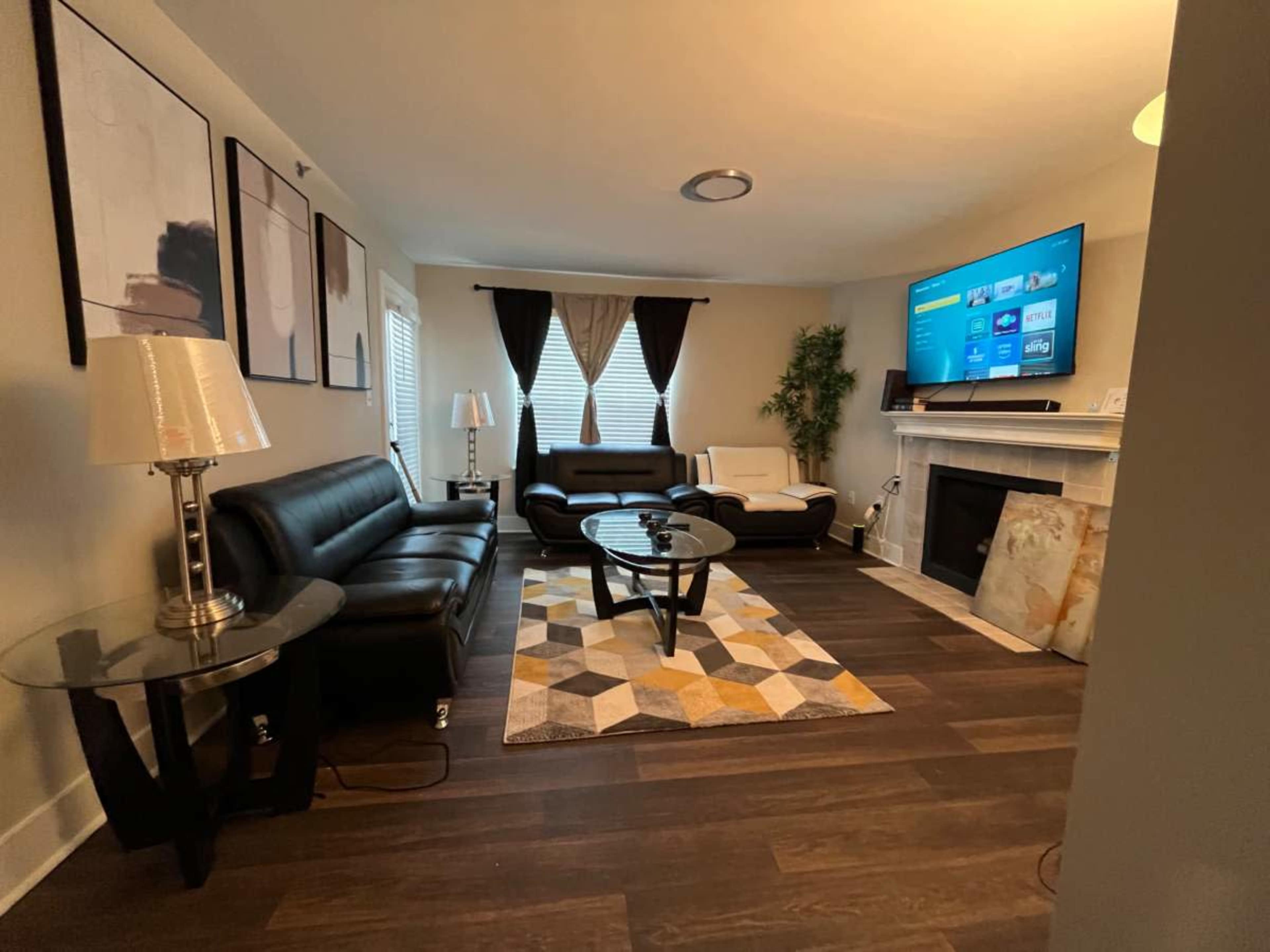 The living room features a black leather sectional couch, a modern coffee table on a geometric rug, and a mounted television above a white fireplace.