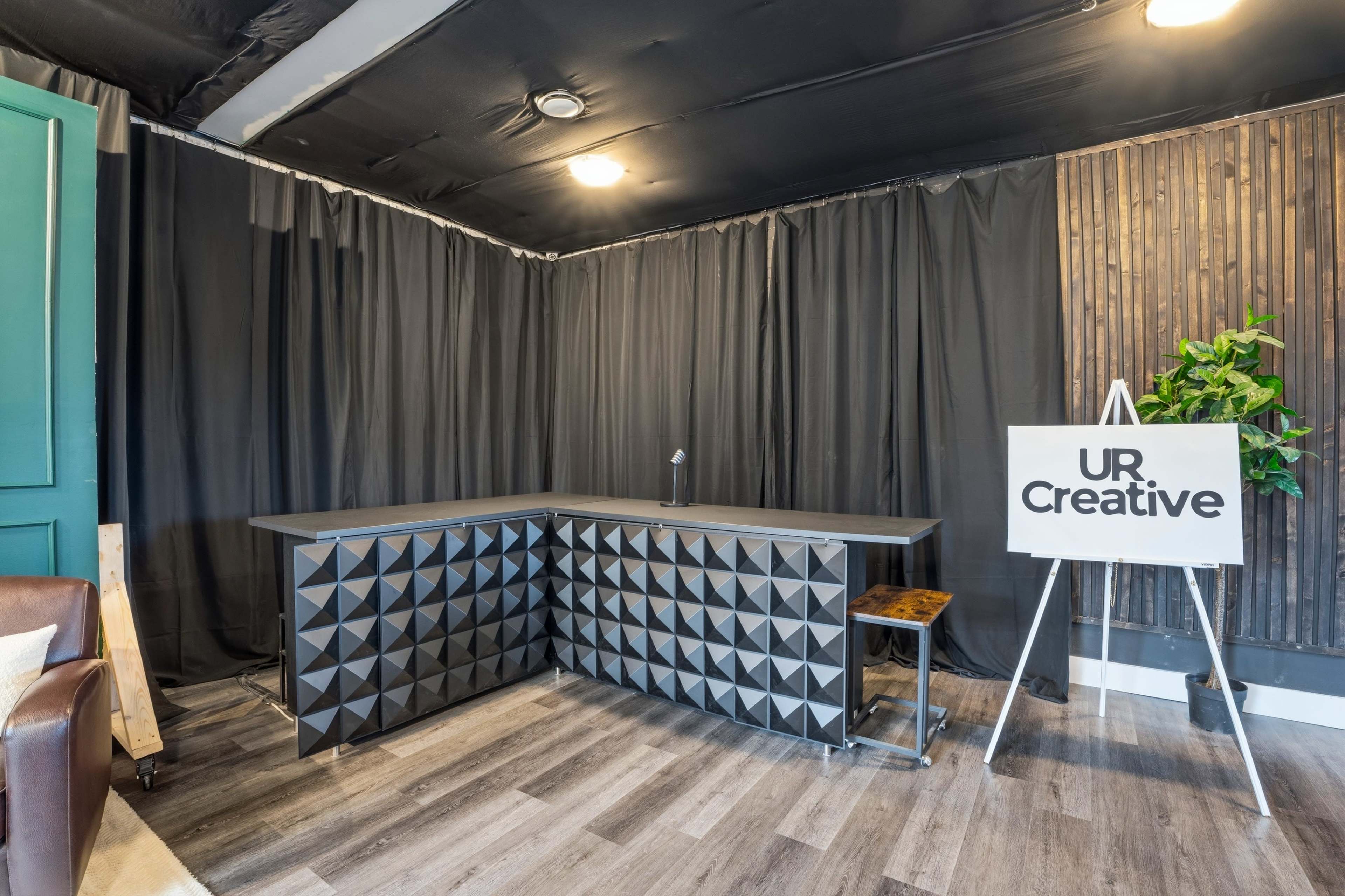 The image shows a modern reception area with a geometric black counter, a sign reading "UR Creative," and a backdrop of black curtains and wooden paneling.