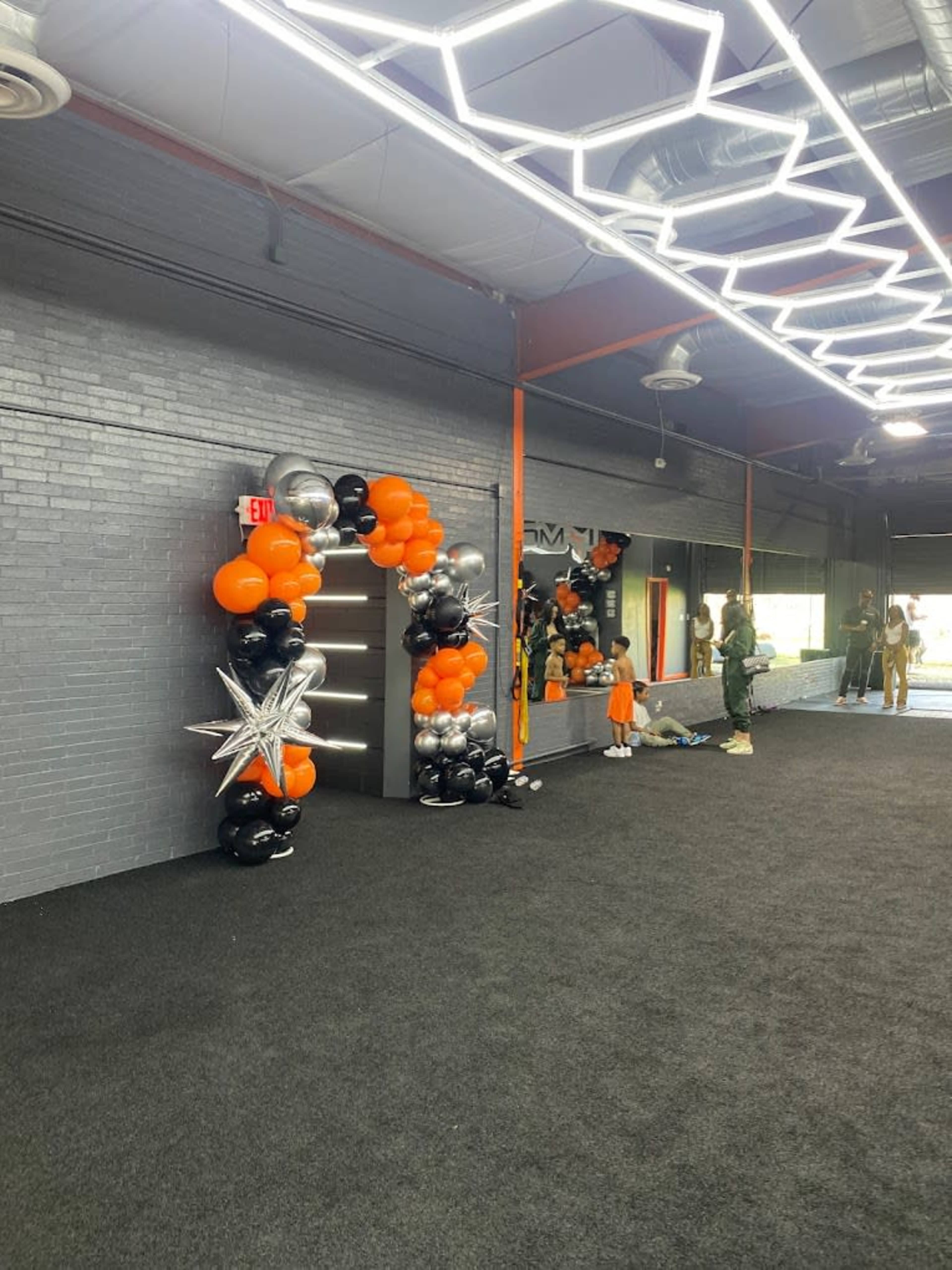 The scene shows a room with black carpeting and a wall decorated with an arch of orange, black, and silver balloons, leading to an exit door.