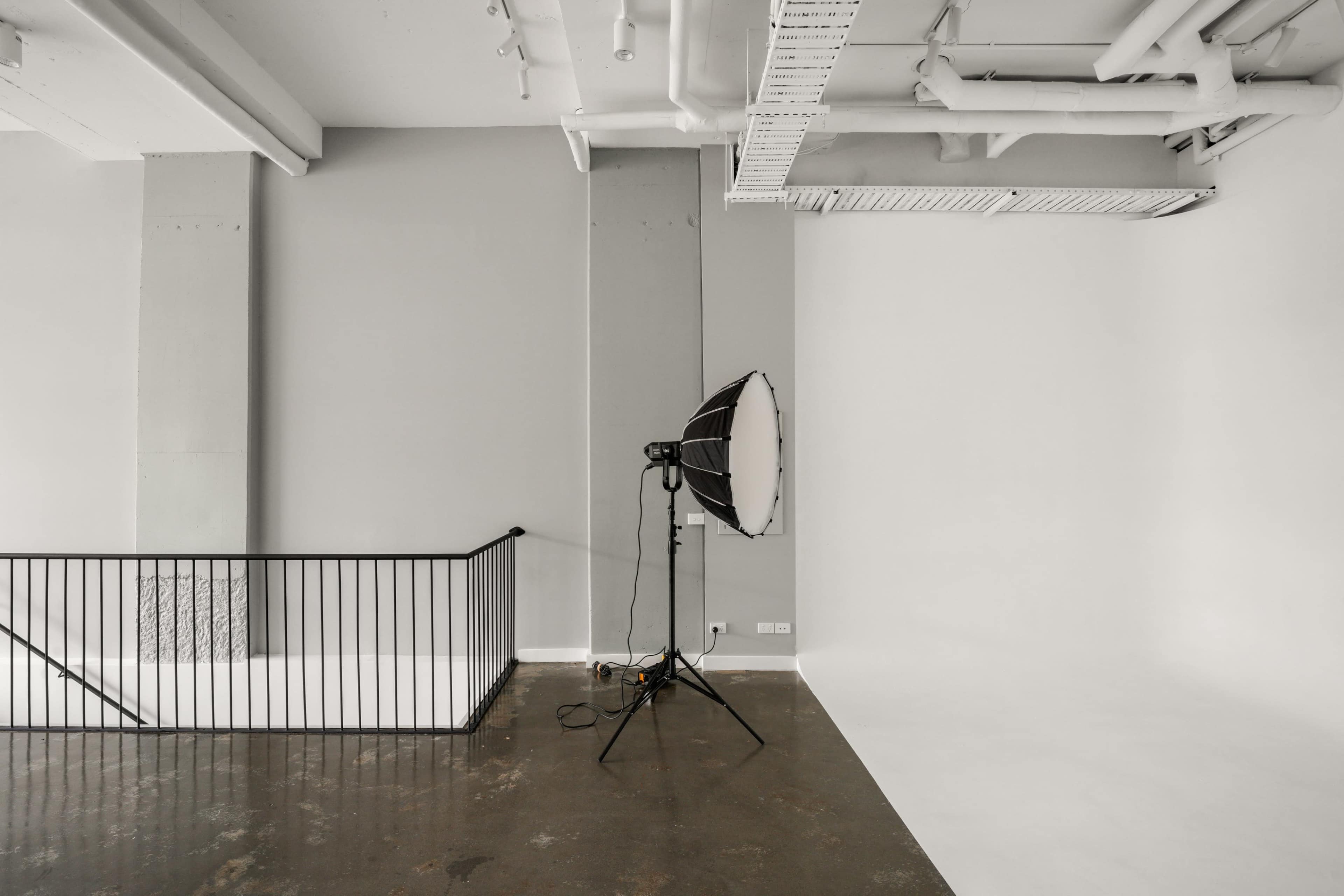 The image shows a minimally furnished photography studio with a large softbox light on a stand and a plain white backdrop.