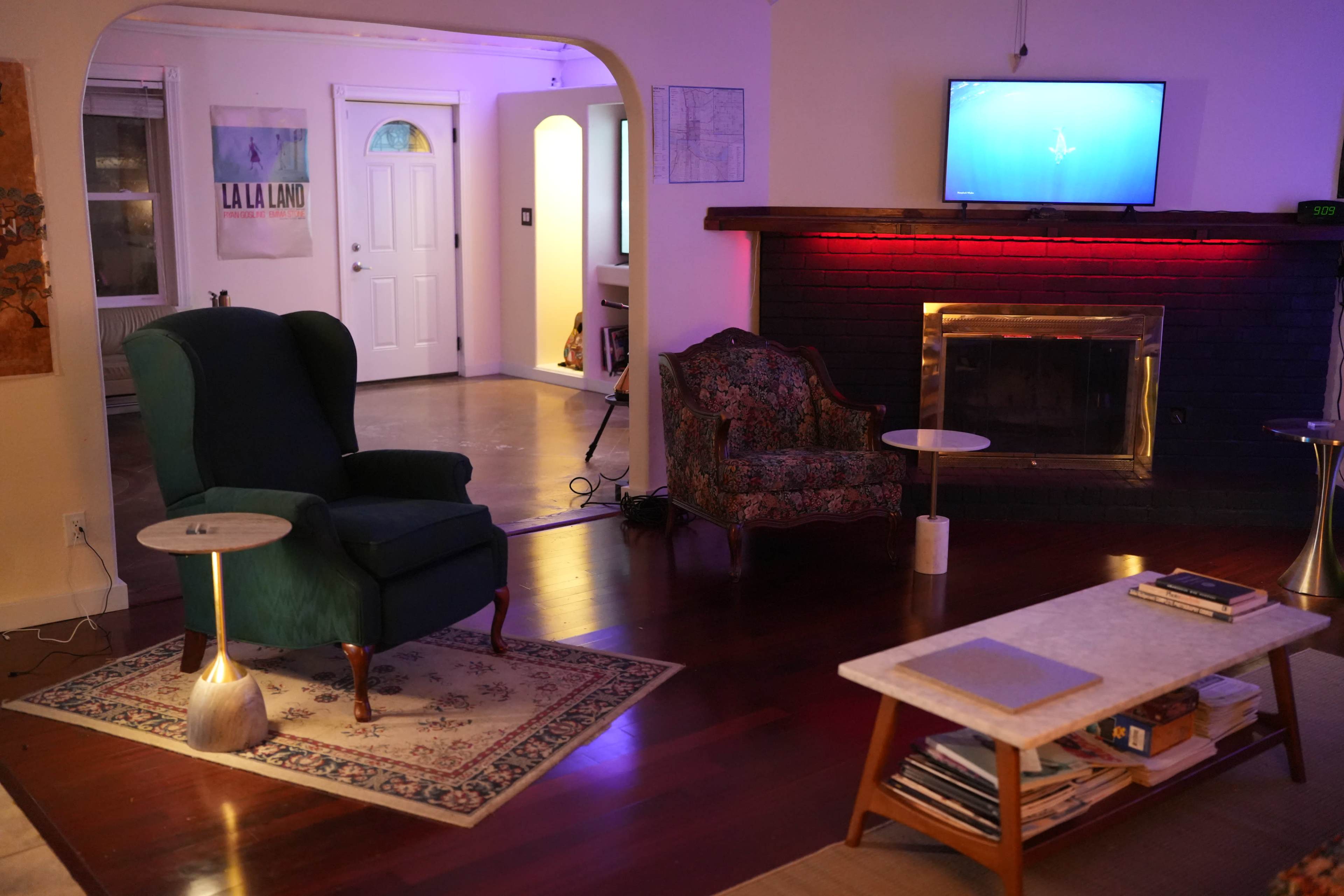 A living room features a green armchair, a patterned loveseat, a coffee table, and a fireplace with colored lighting, all illuminated by soft ambient light.