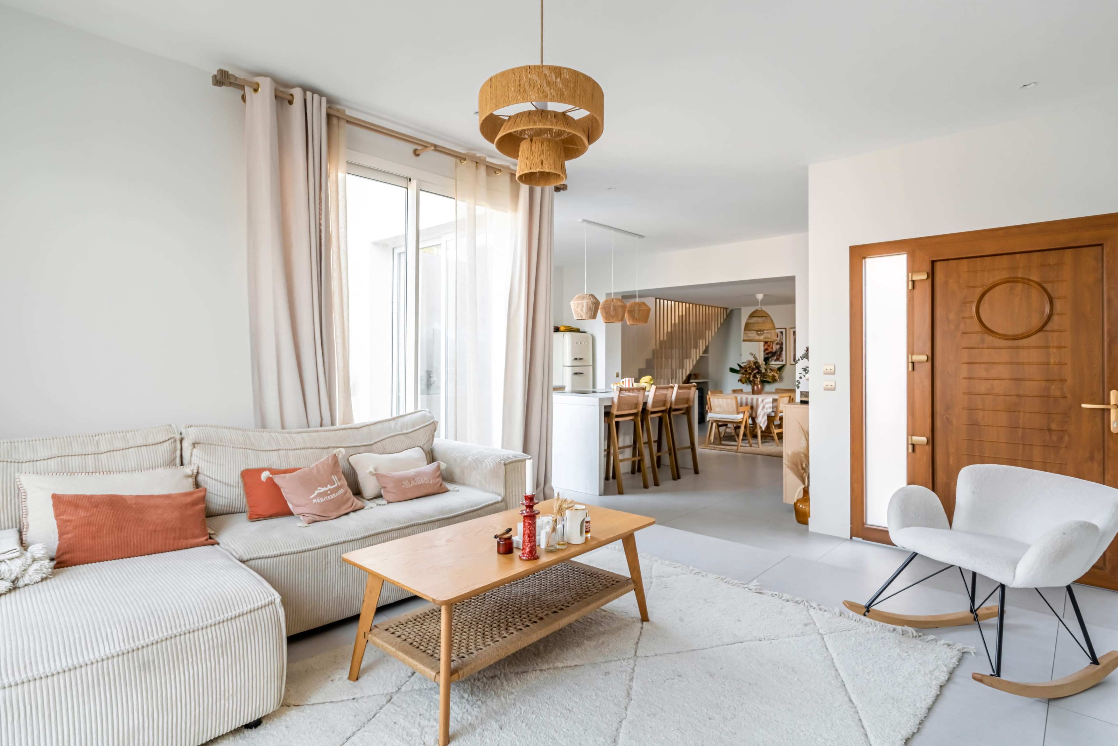 A cozy living room features a light-colored sofa, a wooden coffee table, and a rocking chair, with large windows and a view of an adjoining dining area.