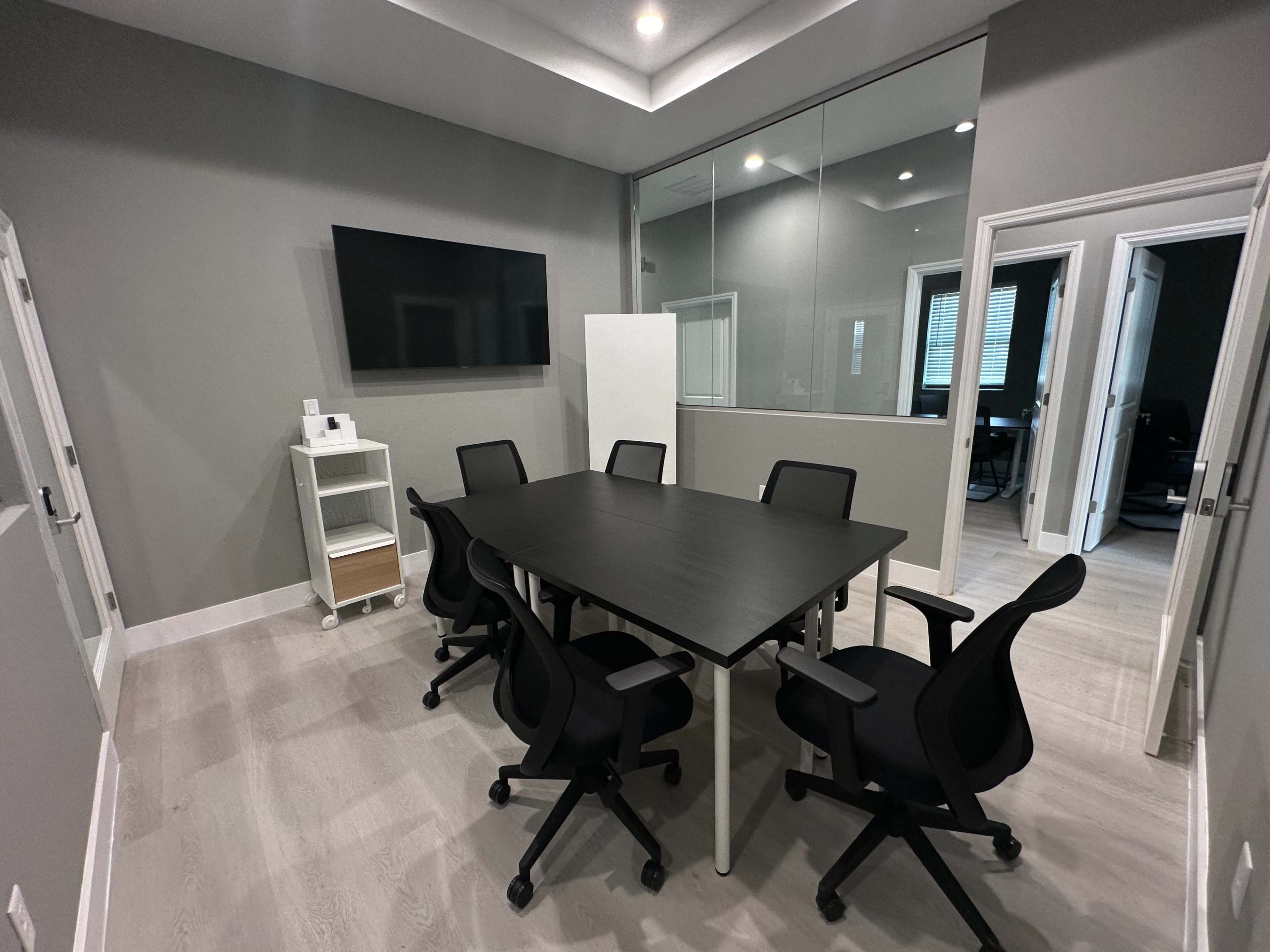 The image shows a modern conference room with a large table surrounded by six black chairs and a wall-mounted television.
