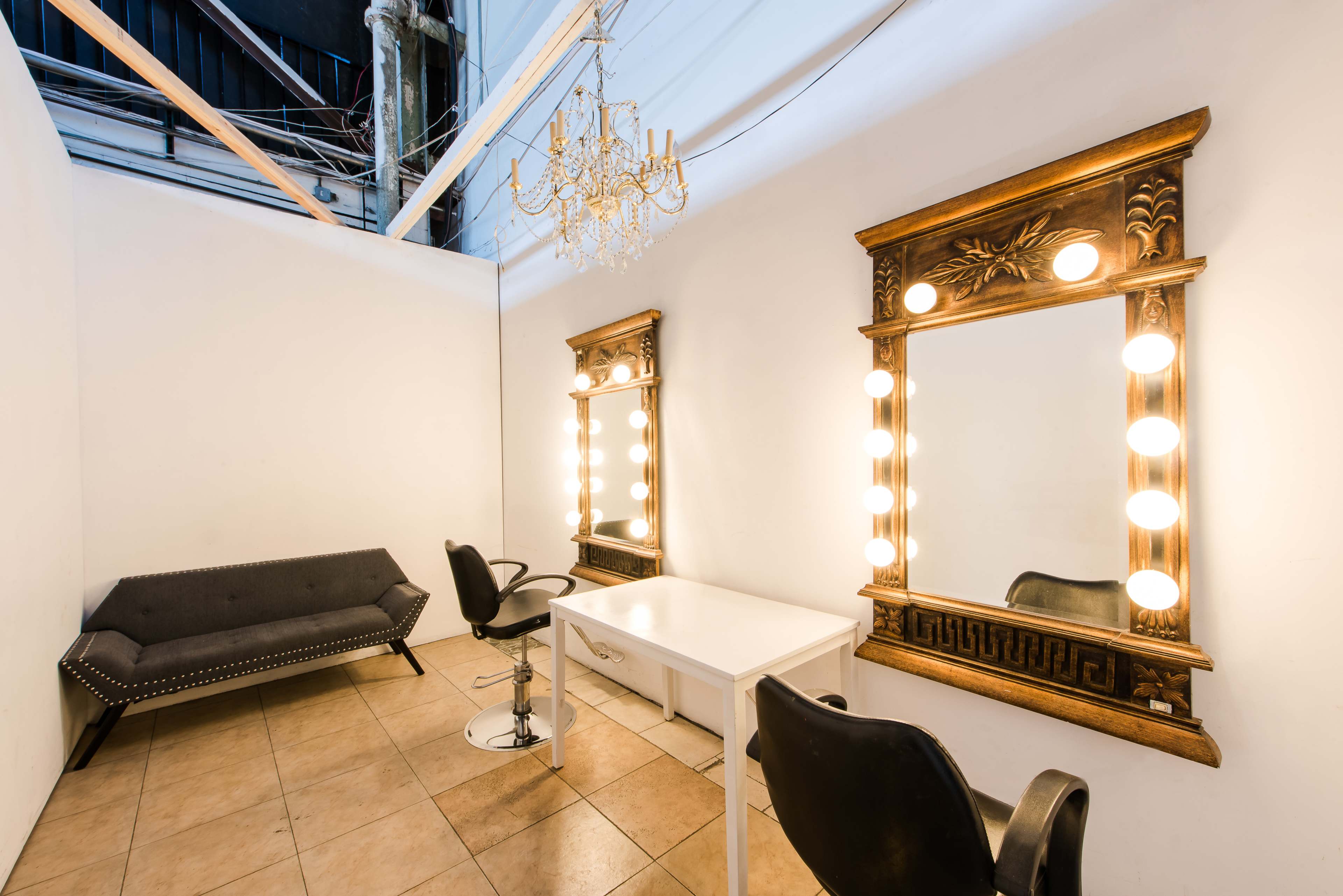 The image shows a well-lit vanity area with two large mirrors surrounded by lights, a small white table, and a black chair, set against a white wall and tile floor.