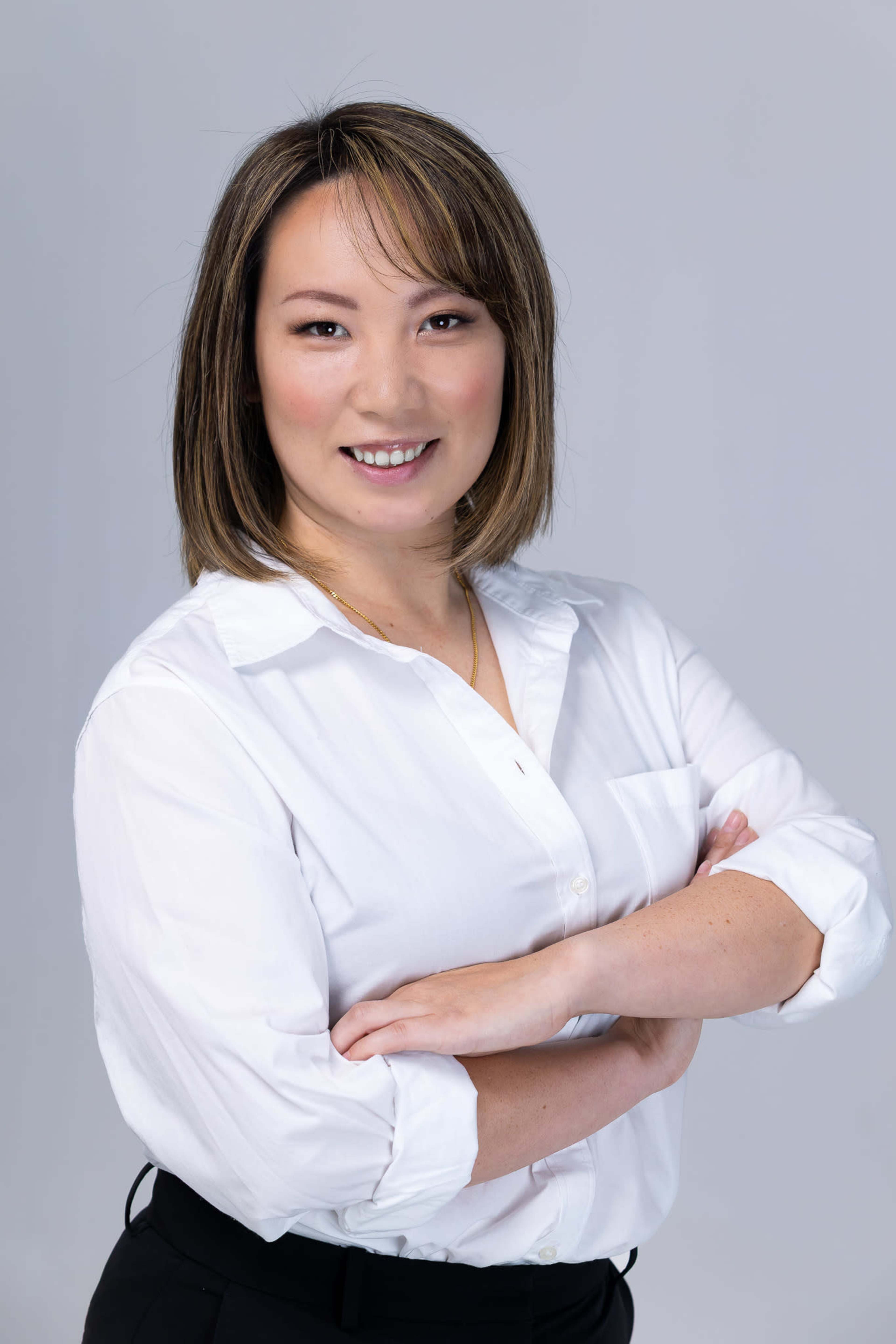 A woman wearing a white button-up shirt stands with her arms crossed, smiling against a neutral gray background.