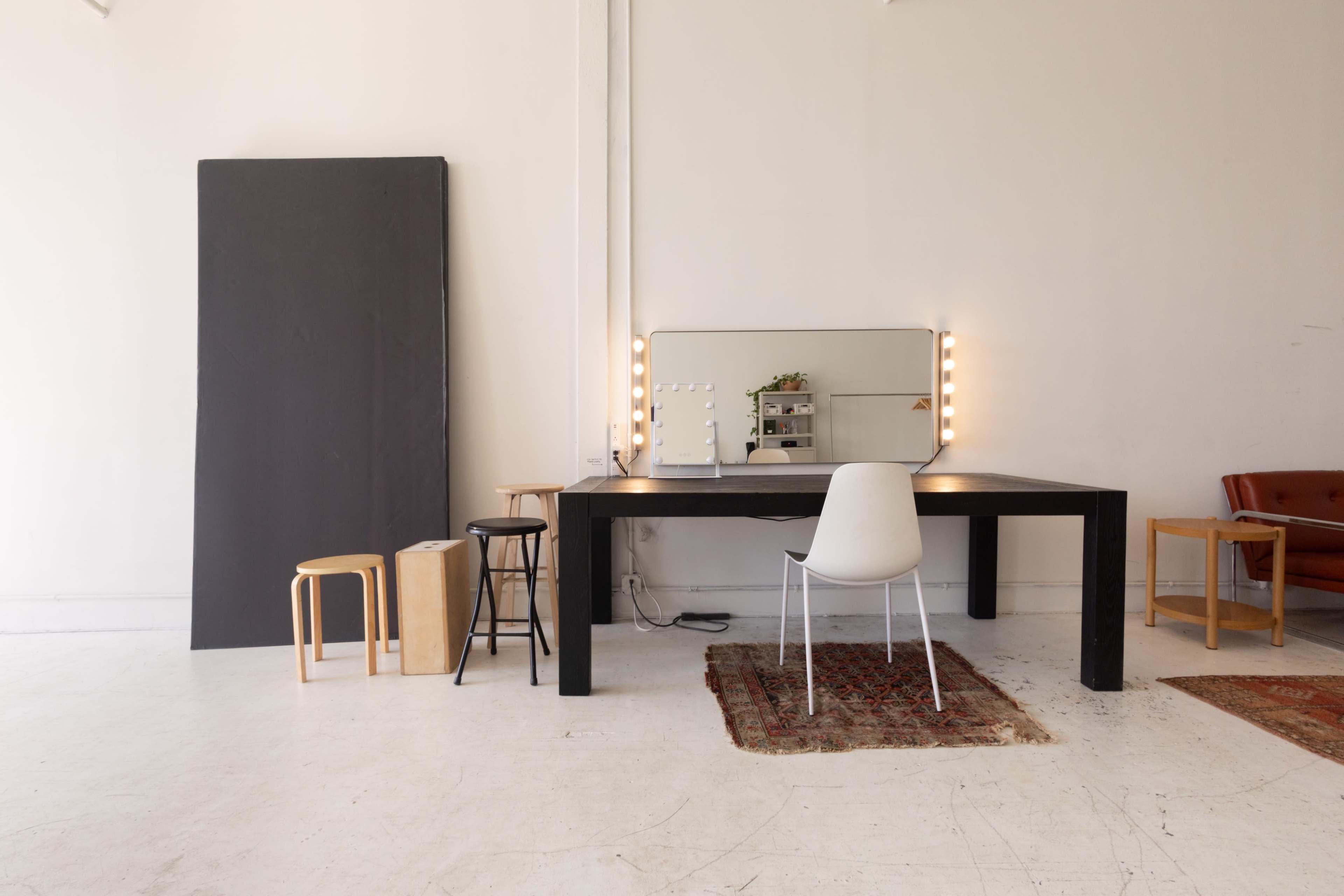 A minimalist room features a large black table with a mirror and light bulbs, surrounded by a mix of stools and a patterned rug.