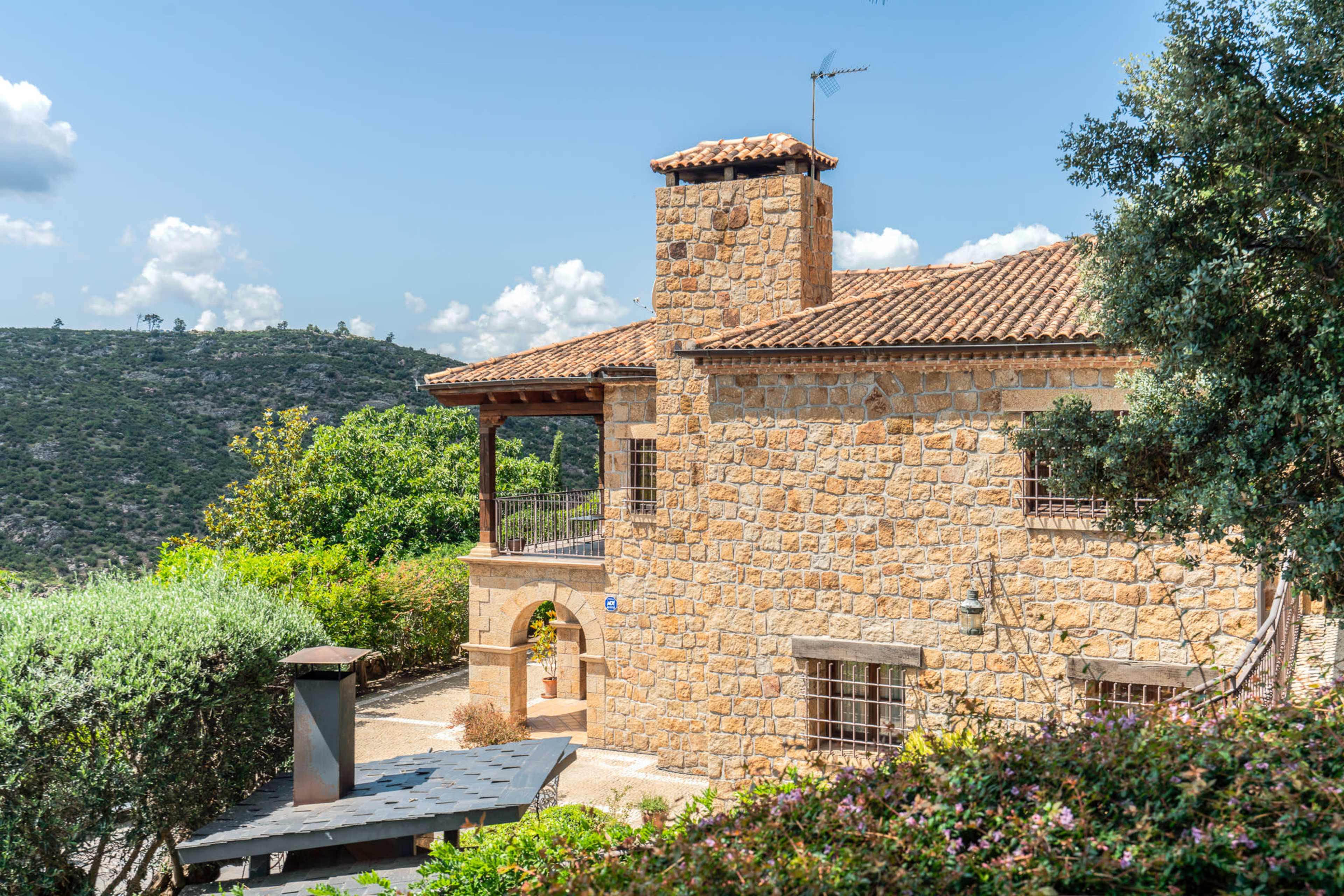 A stone house with a tiled roof and a chimney is set among greenery, overlooking a hilly landscape.