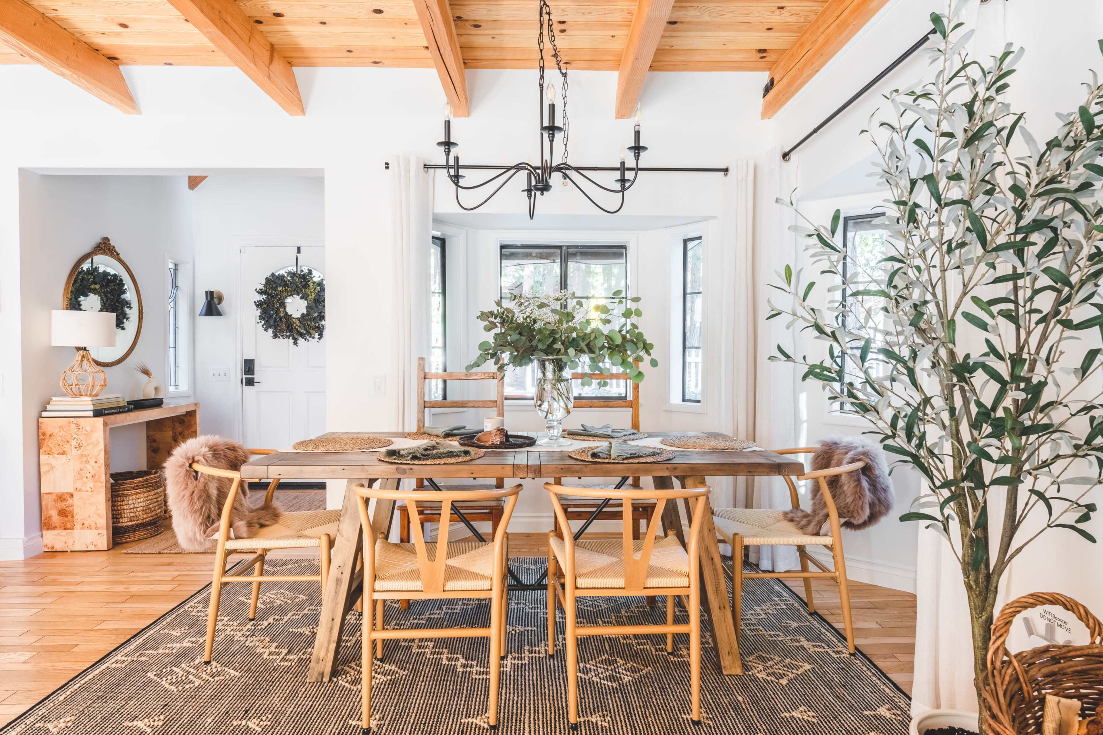 A dining room features a wooden table surrounded by woven chairs, with a chandelier above and large windows on one side.