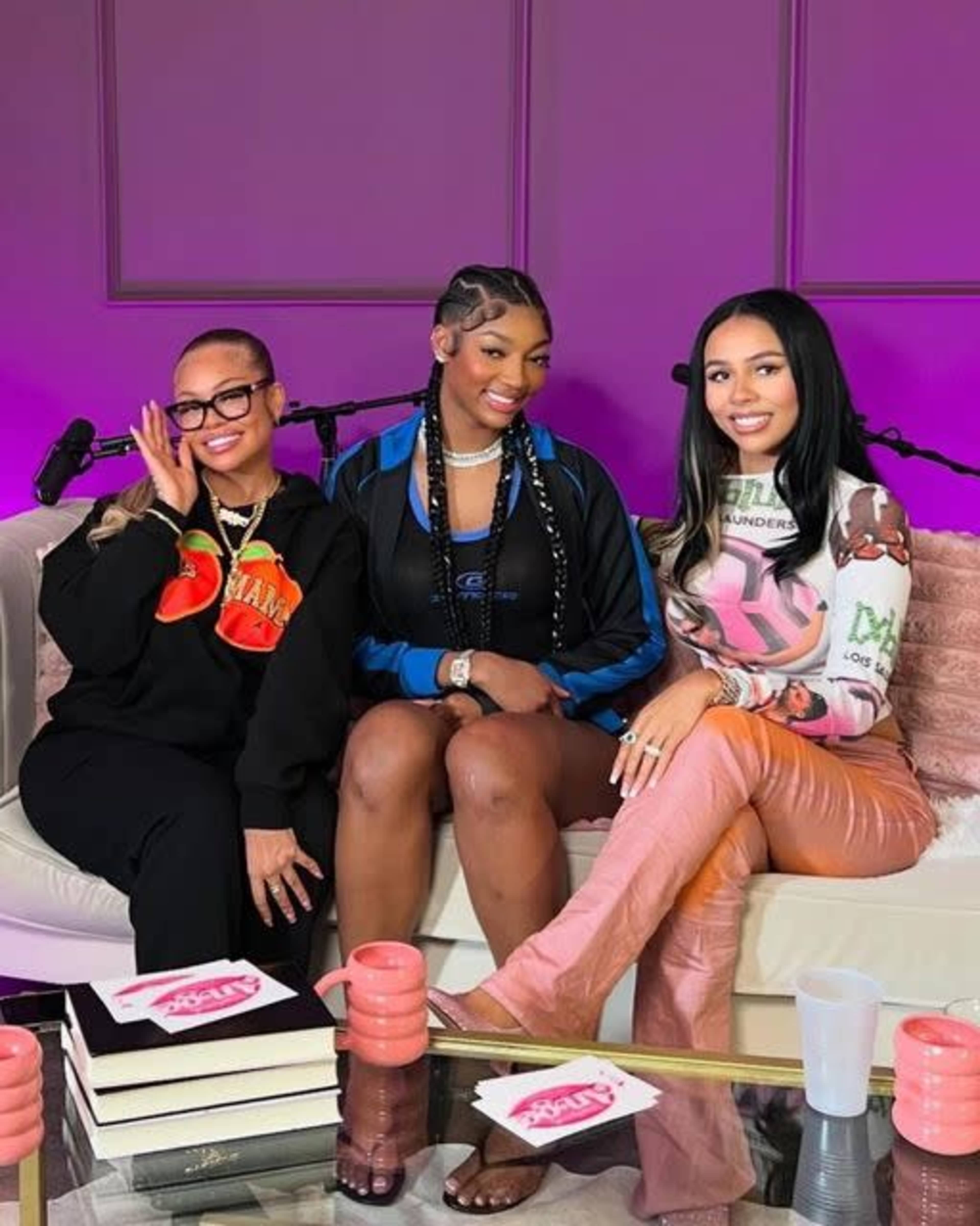 Three women are seated on a couch in a studio setting, dressed in fashionable outfits, with microphones and a pink backdrop in the background.
