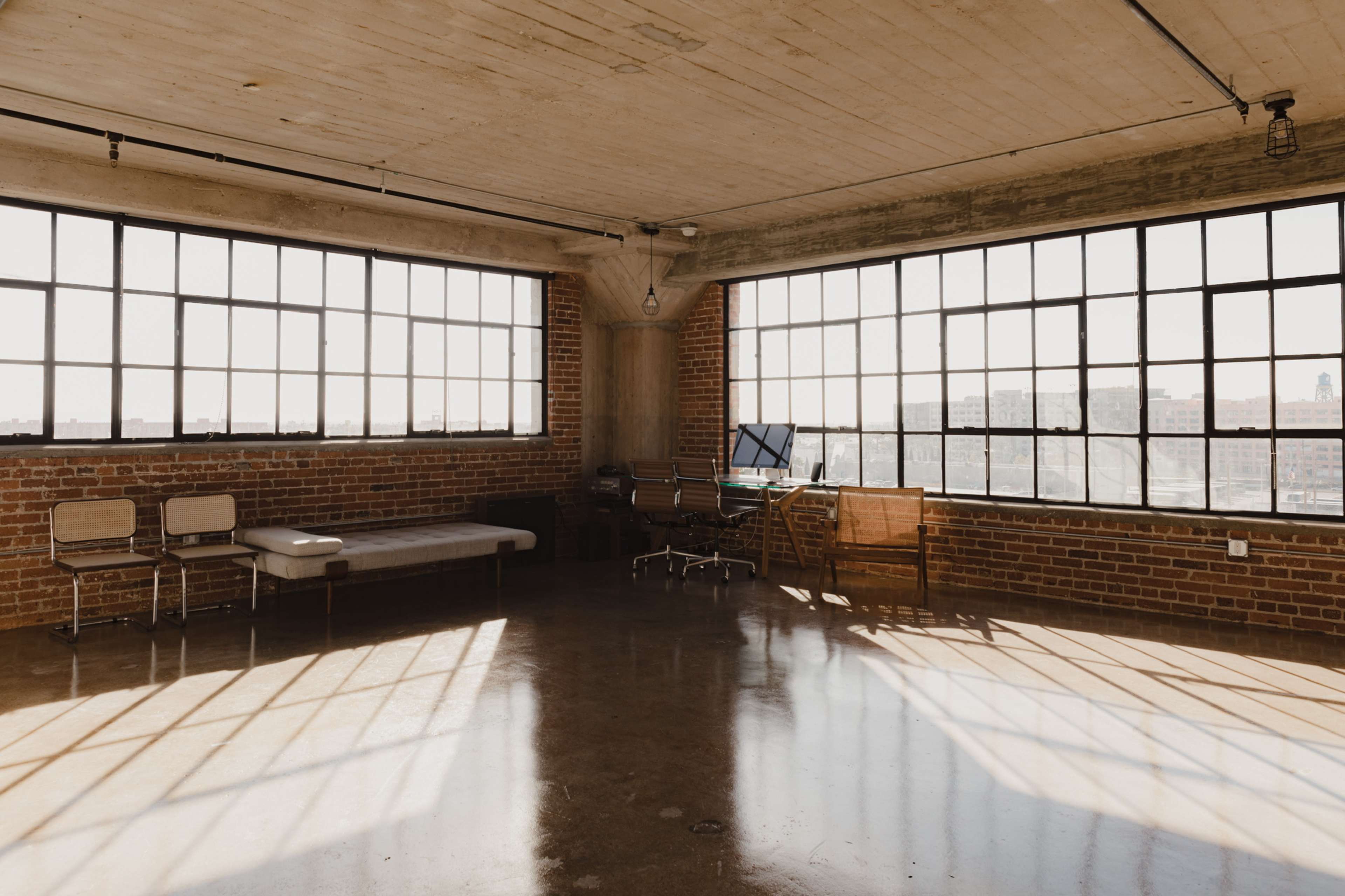 A spacious room with exposed brick walls, large windows, and minimal furniture, including a bed, two chairs, and a desk with a computer.