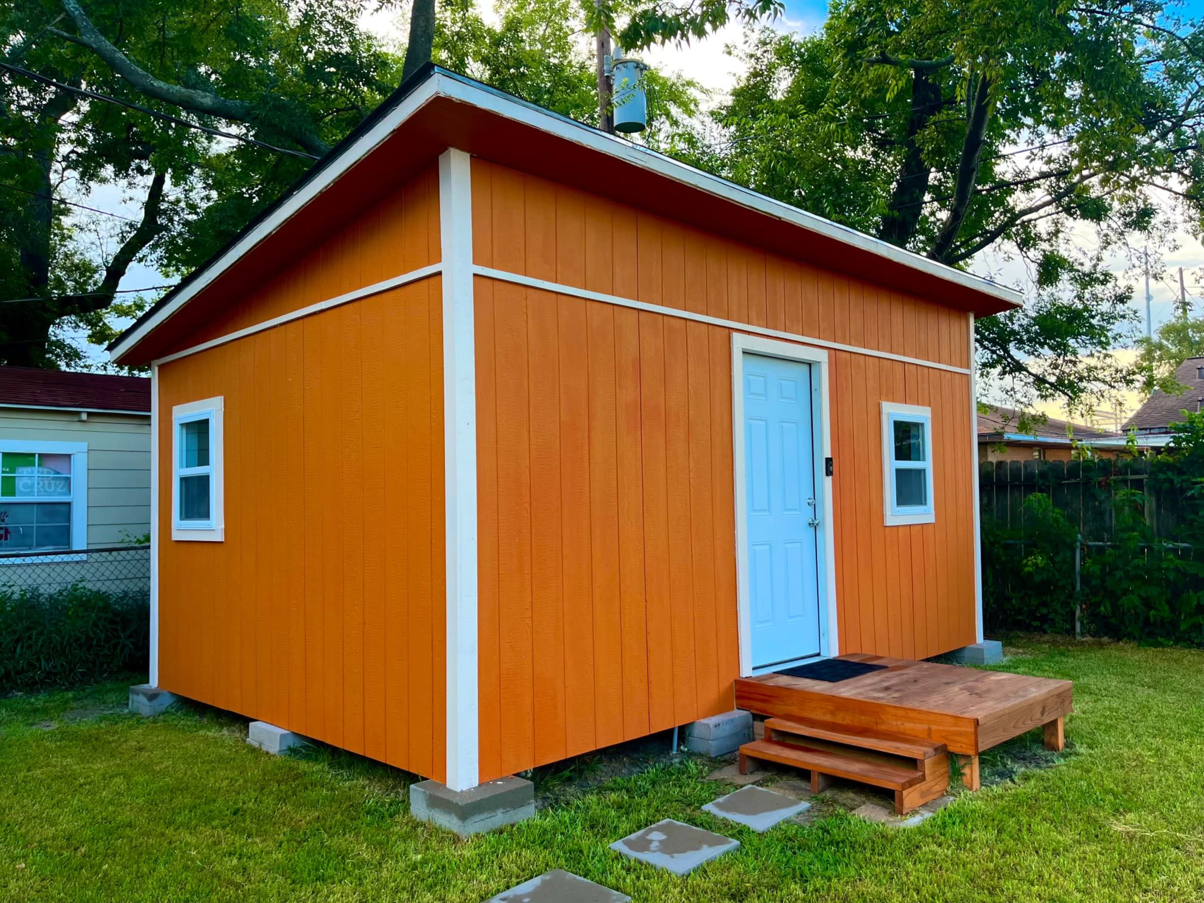 A bright orange shed with a white door and windows is set on a grassy area surrounded by trees and a fence.