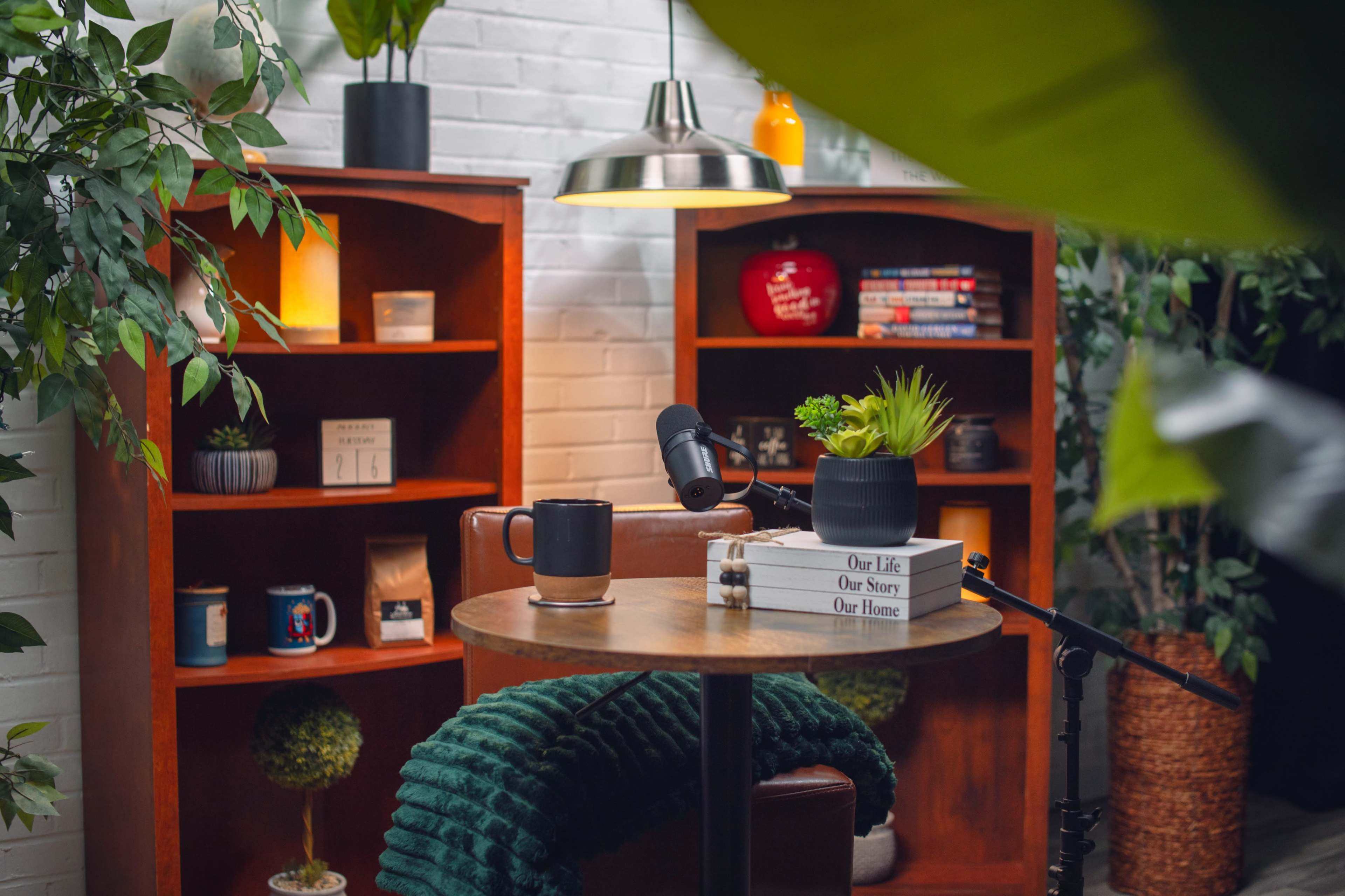 A cozy reading nook features a wooden table with a coffee mug, a potted plant, and a microphone, surrounded by bookshelves filled with plants and decorative items.
