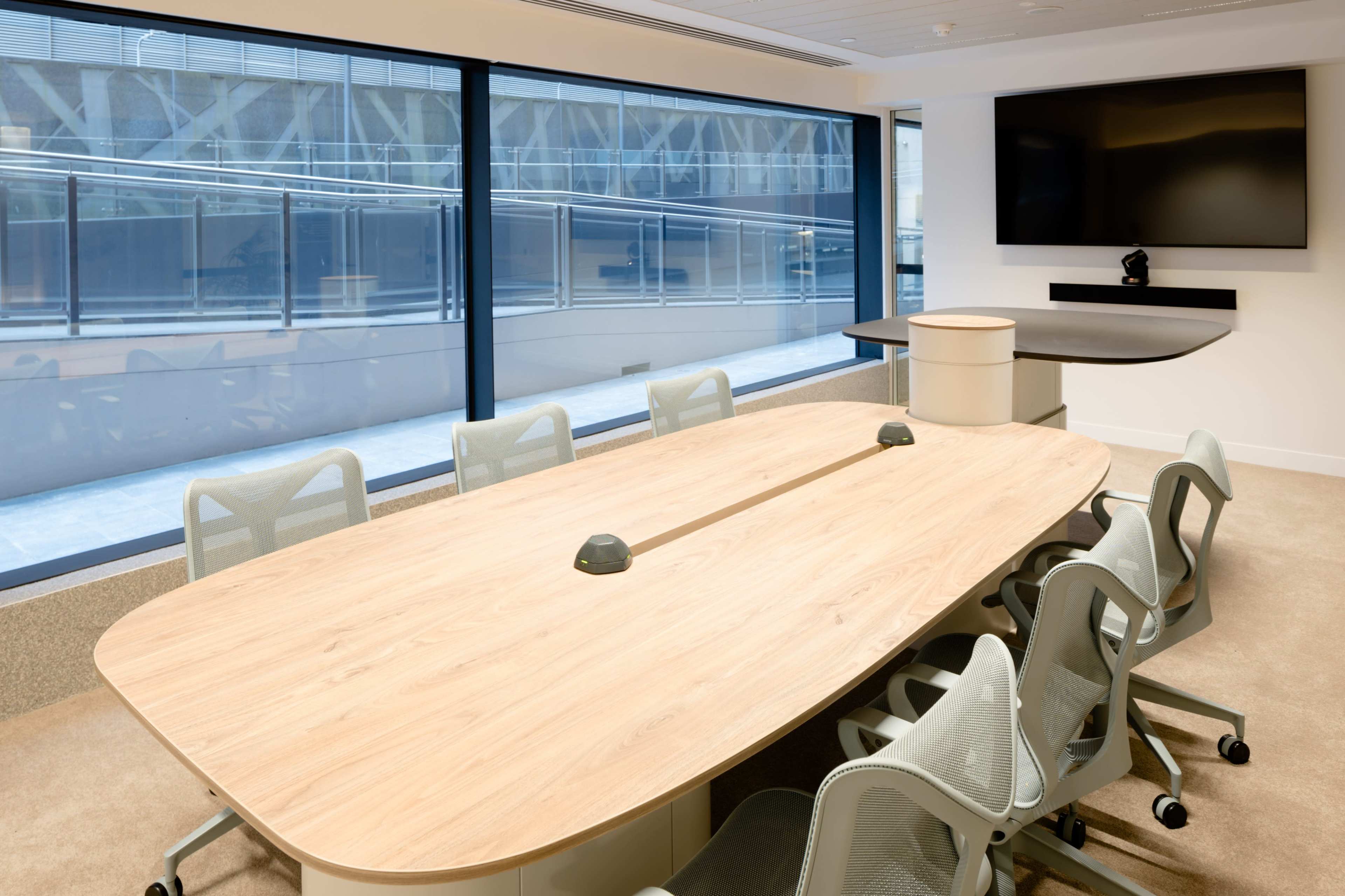 A modern conference room features a large wooden table surrounded by ergonomic chairs and is illuminated by natural light from large windows.