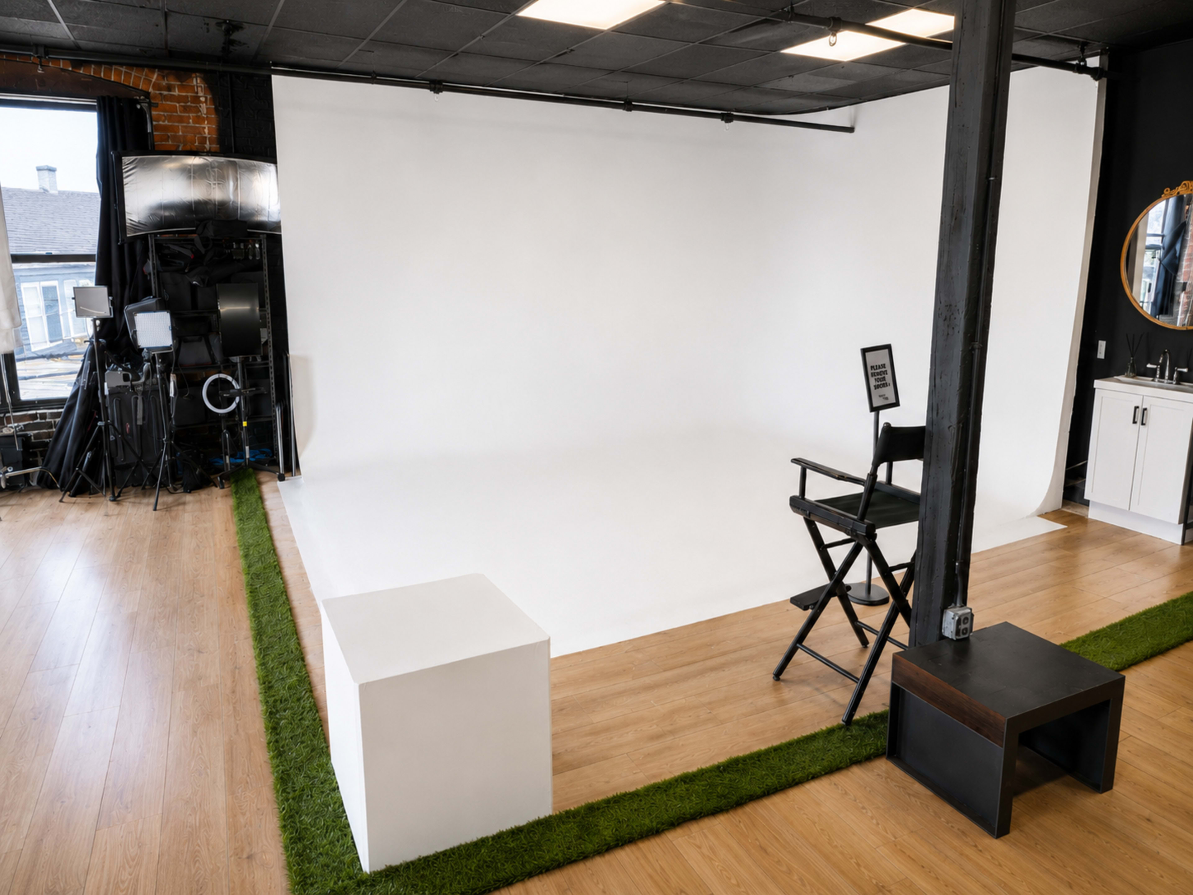 The image shows a minimalistic photo studio featuring a white backdrop, a director's chair, a side table, and a mirror, with wooden flooring and green grass-like carpet accents.