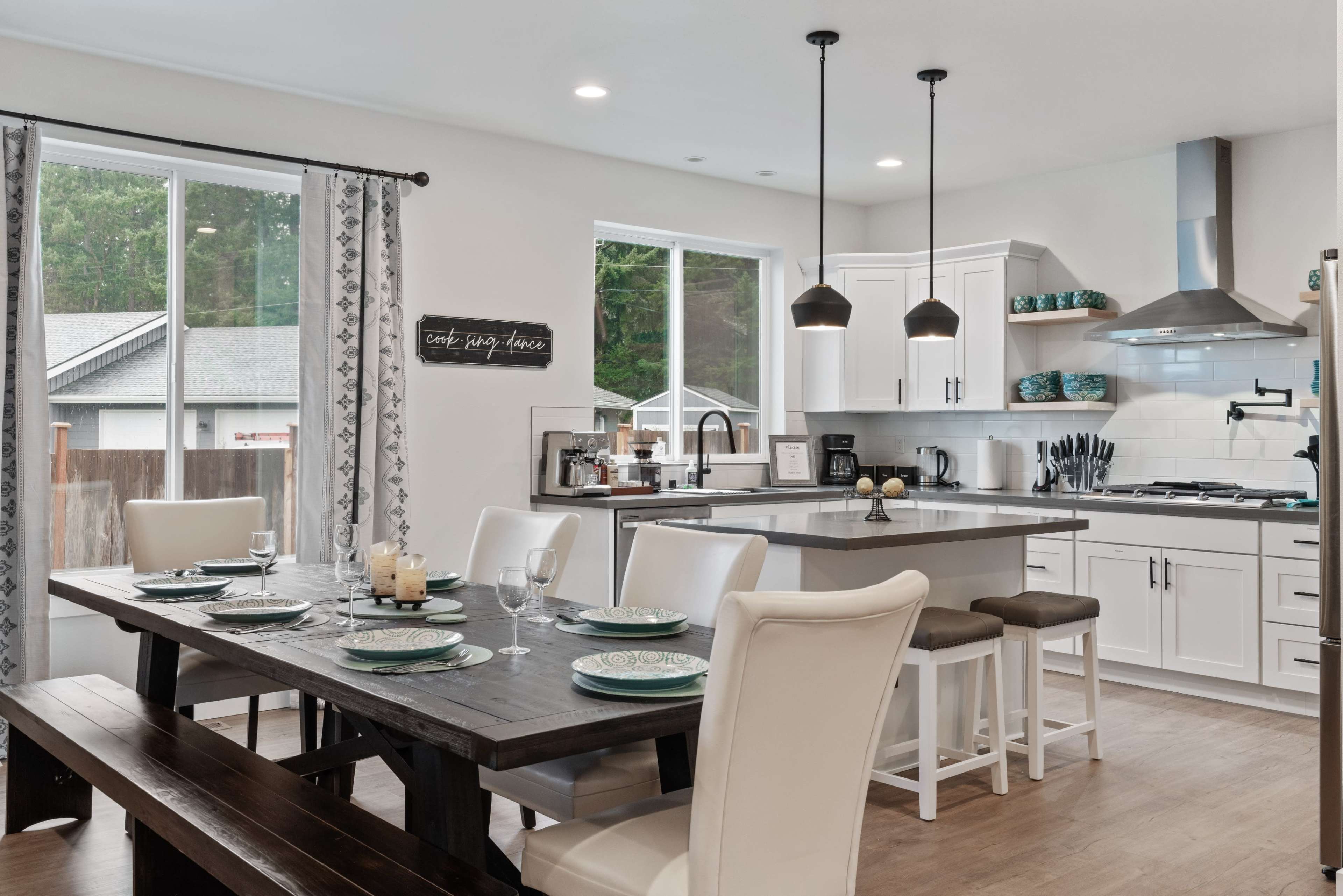 A modern kitchen and dining area featuring a wooden table set for dinner, with white cabinetry and large windows providing natural light.