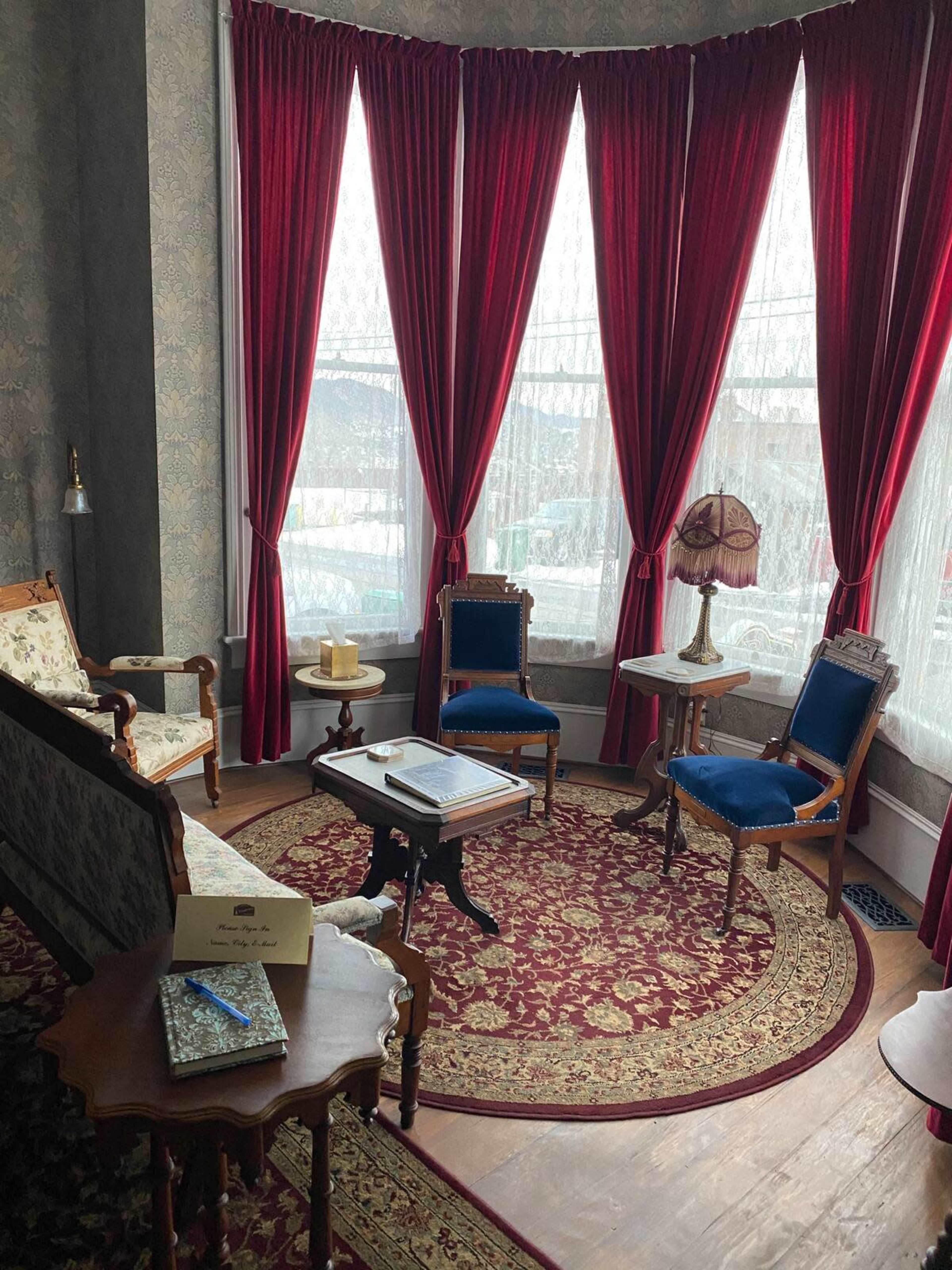 A corner of a room with a patterned rug, vintage furniture, and large red curtains covering a bay window.
