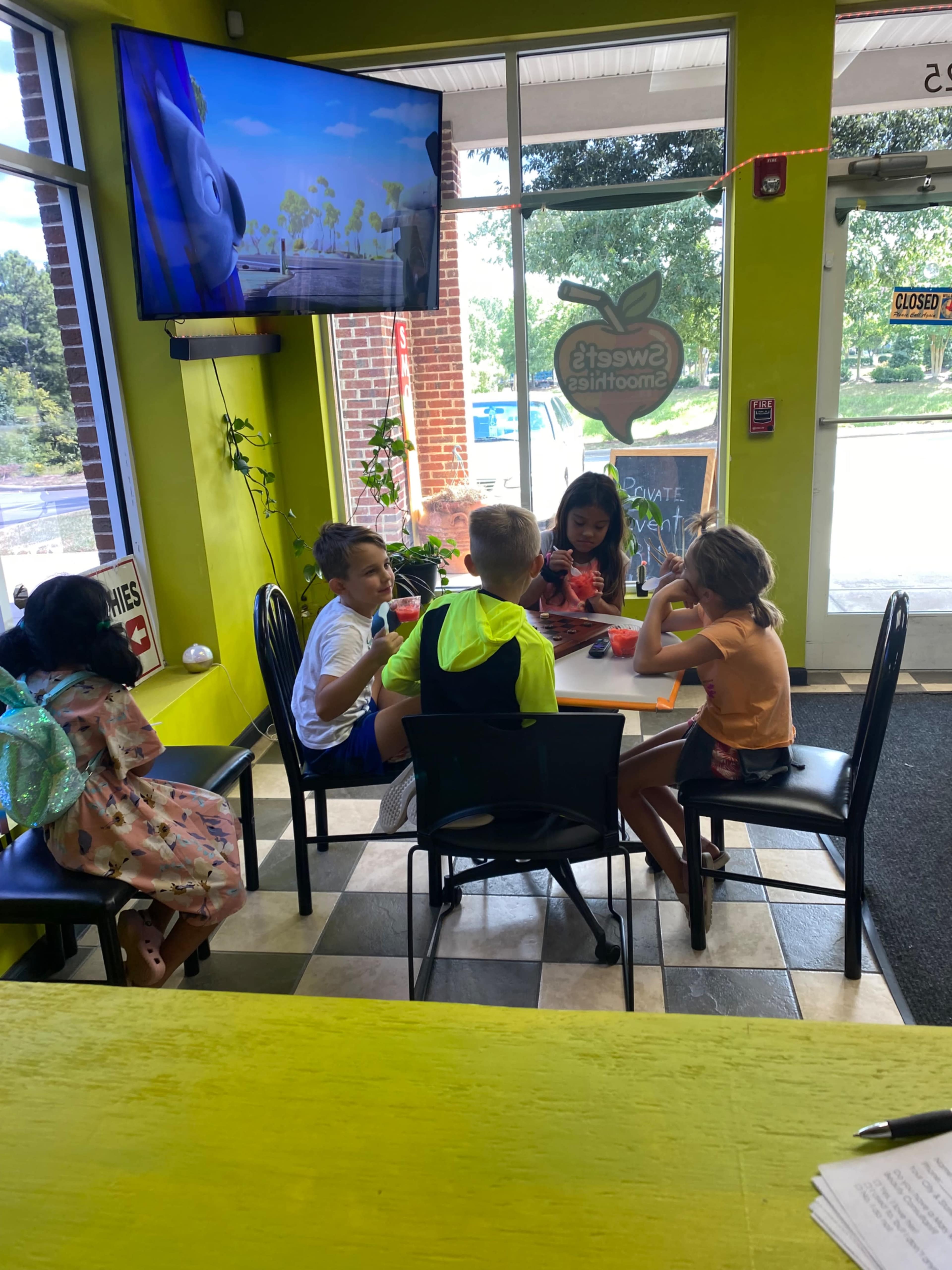 A group of children sits around a table in a bright room, enjoying snacks while watching a cartoon on a television.