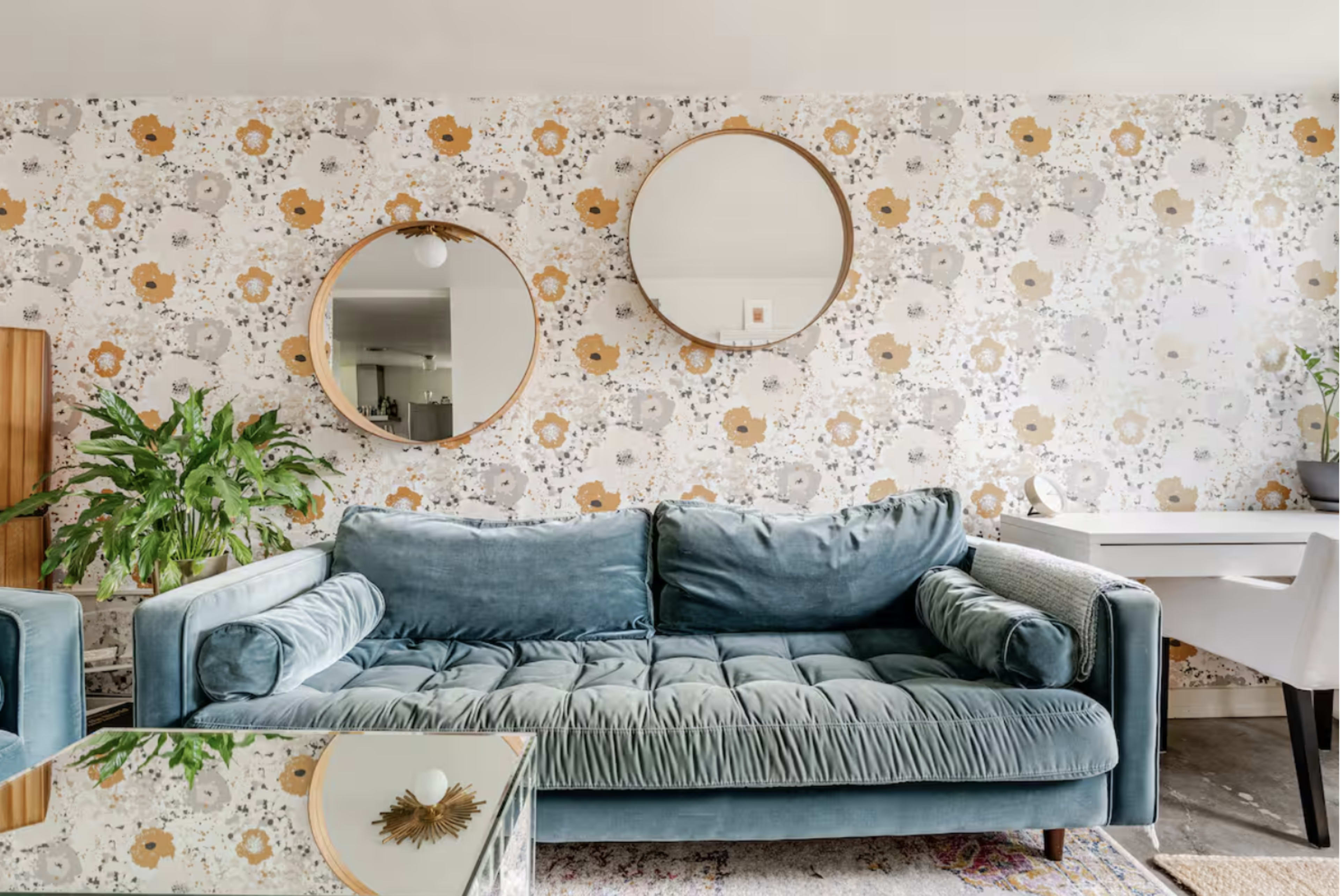 The image shows a living room with a blue velvet sofa, two round mirrors mounted on a floral-patterned wallpaper, a potted plant, and a glass coffee table.