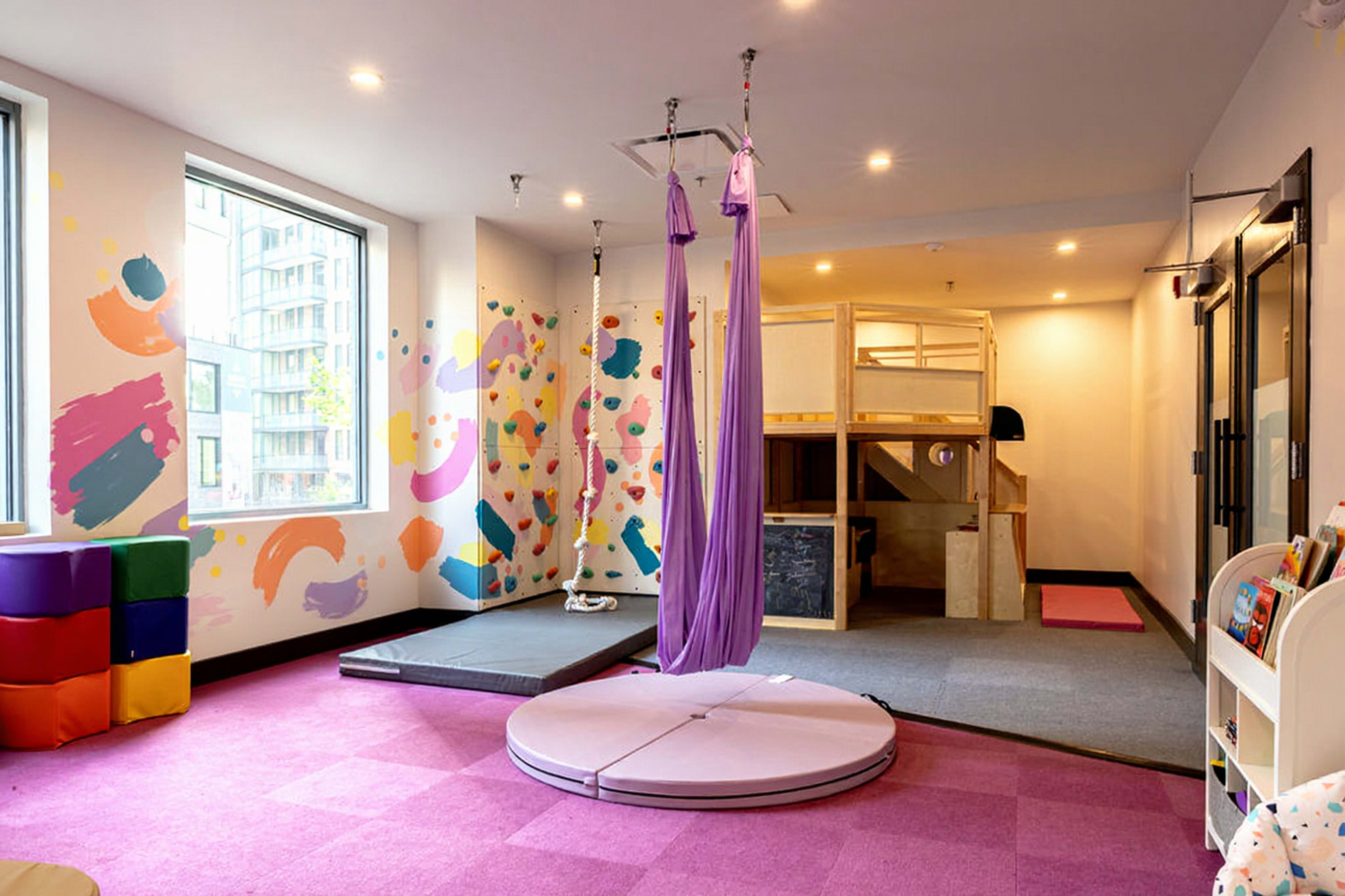 The image shows a brightly colored playroom with a purple mat, a circular padded area, a climbing structure, and suspended fabric for play.